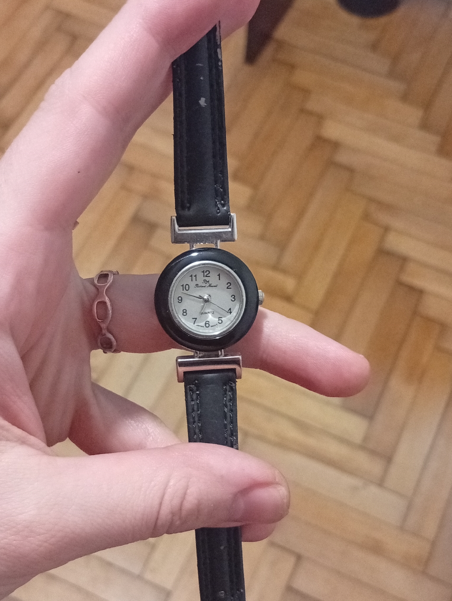 A person's hand holds a black leather strap watch with a round face. The watch face has white numerals and black trim. The watch brand name is "Richard Morel" and the dial indicates it is a Quartz movement. The background consists of a wood floor. The location in the city of Buenos Aires, Argentina is indeterminate from the provided image.