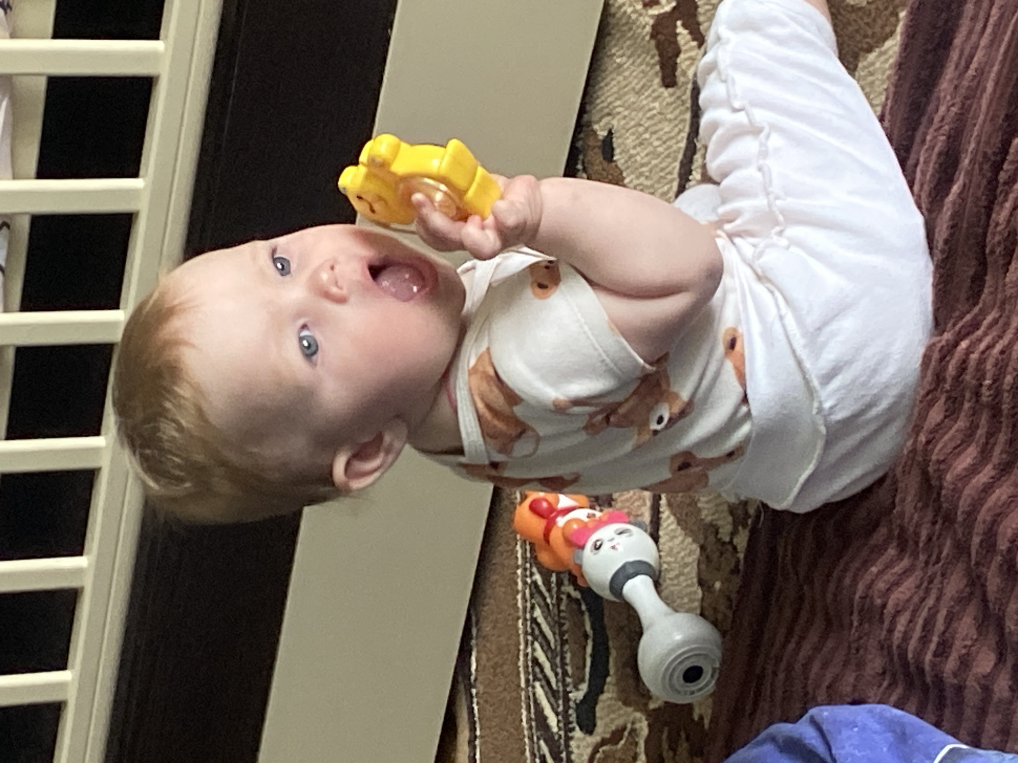 A baby with light hair and blue eyes is lying on a patterned carpet and a brown textured blanket. The baby is wearing white pants and a white shirt with a brown deer pattern. The baby holds a yellow toy in their right hand. Next to the baby is a baby monitor and part of a crib is visible in the upper left. The location is most likely a home, but it is not possible to confirm the location in Фоки, Russia.