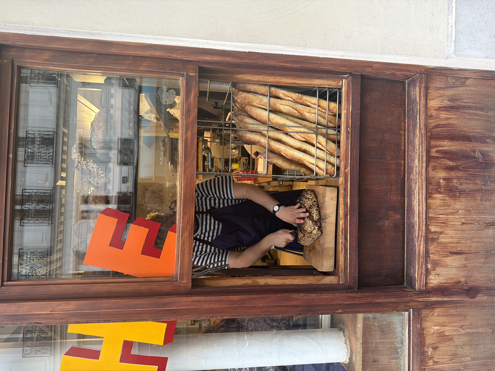 A wooden-framed storefront window in Paris, France. Inside the window, baguettes are stored in a metal basket. A person wearing a striped shirt and blue apron slices a loaf of seedy bread on a wooden cutting board. The window also displays colorful signage, and the reflection shows buildings across the street with wrought iron balconies.