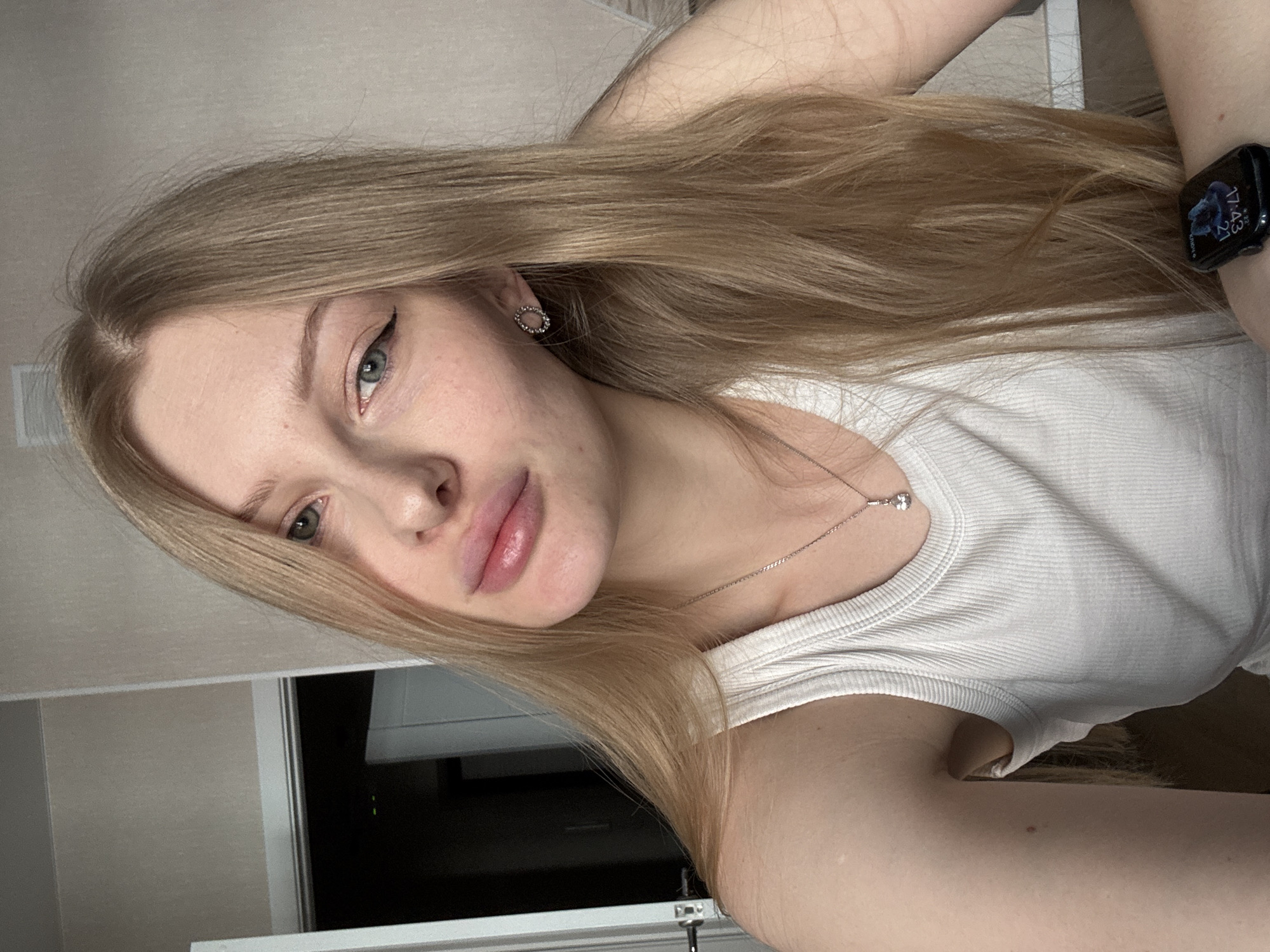 A fair-skinned young woman with long, light-blonde hair is seen in a selfie. She is wearing a white tank top and a silver necklace with a small pendant. Her makeup includes winged eyeliner and lip color. She has a stud earring. A smart watch is visible on her wrist, displaying a clock face with a time of 17:43 and a date of 27. The background includes a door and a light-colored wall. There is no evidence in the image to suggest the subject is in Perm, Russia.