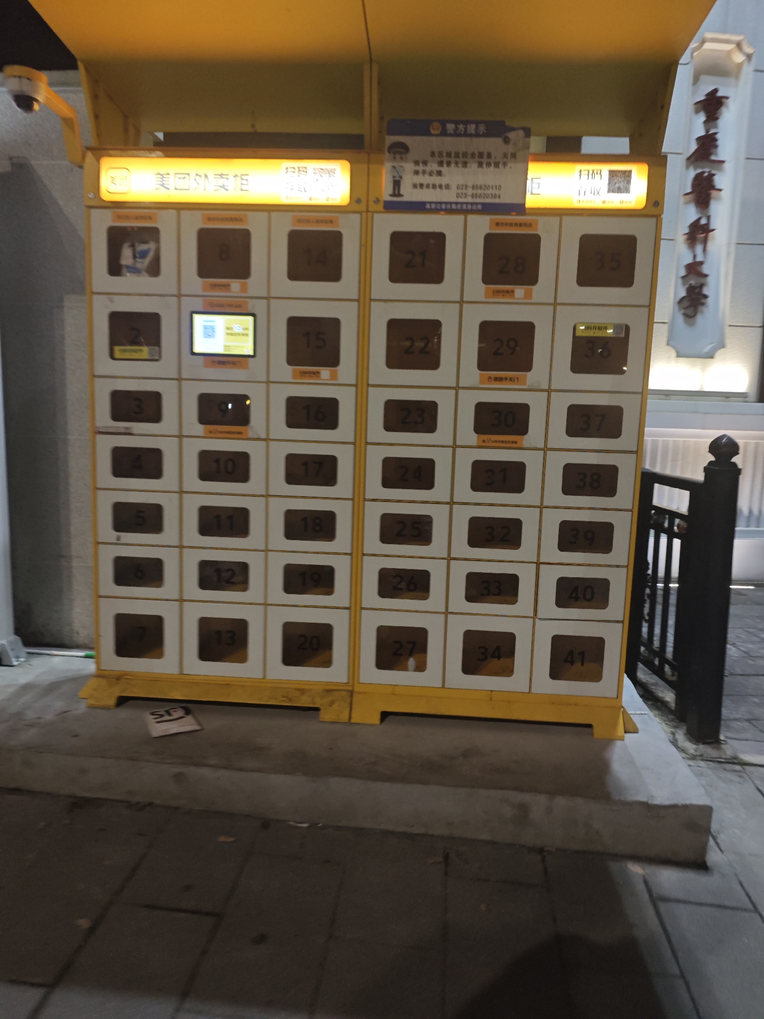 The image shows two delivery lockers, likely for food delivery services, situated outdoors. They are located in the Chongqing High-tech Zone, China, as indicated by the Chinese characters on a nearby sign. The lockers are painted yellow and white, with numerical designations from 1 to 41 on individual compartments. The sign on the top of the left locker contains the Chinese characters "美团外卖柜" (Měituán wàimài guì), which translates to "Meituan Takeout Locker." The right locker contains an information notice with a telephone number for help. Next to the right locker is a black metal fence, suggesting a street-side location. The environment appears to be urban, with pavement visible in the foreground and building structures visible in the background.