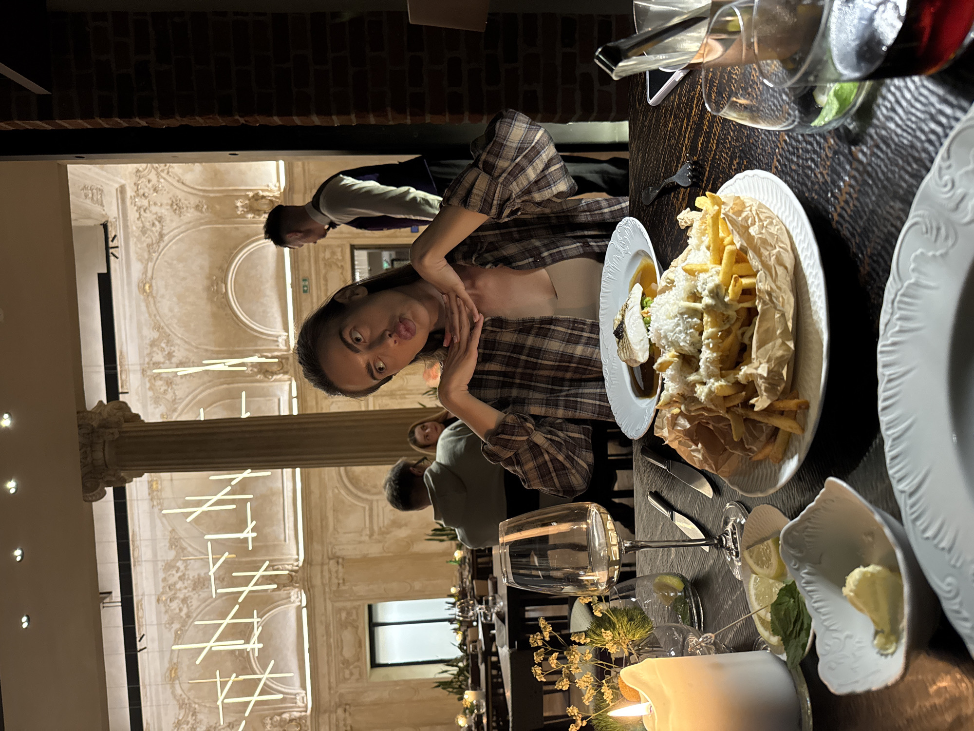 The image depicts an indoor scene at an upscale restaurant located in Unknown, Unknown. In the foreground, a young woman with long dark hair, wearing a plaid shirt over a light-colored top, is seated at a dark wooden table. She is looking upwards and to her left with a puckered lips expression, and her hands are clasped under her chin in a playful pose. On the table in front of her are several dishes: a white plate holding a serving of french fries in brown paper, topped with a white sauce or cheese; another white plate with a piece of fish on a bed of vegetables with a brown sauce; a small white dish containing lemon wedges and a green herb; and another small white dish with a pat of butter. Various glasses, including a wine glass, a glass with red liquid, and another with a green garnish, are also on the table. A lit white pillar candle and a small floral arrangement are present as table decor. In the mid-ground, other patrons are visible: a man in a grey hoodie seated at a table to the left, and a woman with reddish hair partially obscured behind him. To the right, a male waiter, dressed in a white shirt, dark vest, and dark trousers, stands with his back to the camera near a doorway or partition. The background features high ceilings, ornate classical architecture with columns, arches, and decorative moldings, and modern linear light fixtures hanging from above. A brick wall is visible on the far right.