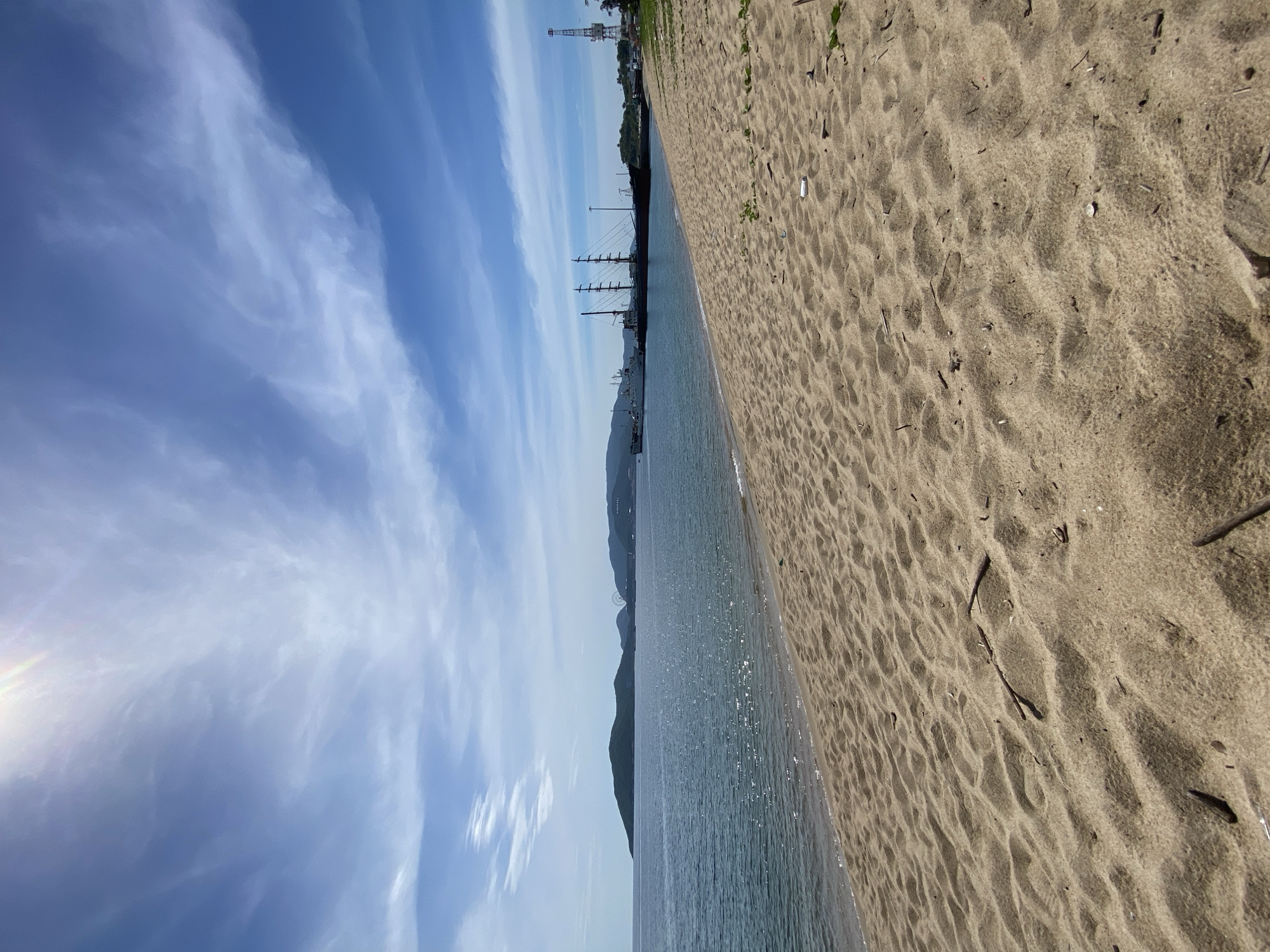 The image depicts a scenic view of a sandy beach and the ocean, likely in Nha Trang, Vietnam. The foreground is dominated by the textured sand of the beach, with small pieces of driftwood scattered across its surface. In the middle ground, the calm ocean stretches out, reflecting the bright sunlight, creating a shimmering effect. Further in the distance, a landmass with hills and mountains can be seen, along with several docked ships. One ship, in particular, stands out with its multiple masts, suggesting it might be a sailing vessel or a historical ship. A tall crane is also visible on the right side of the image, indicating industrial activity or port infrastructure. The sky above is a vibrant blue, adorned with wispy white clouds, and a bright sun is positioned in the upper left corner, casting a lens flare. The overall scene conveys a sense of tranquility and coastal beauty, potentially a popular tourist destination or a less-developed stretch of coastline.