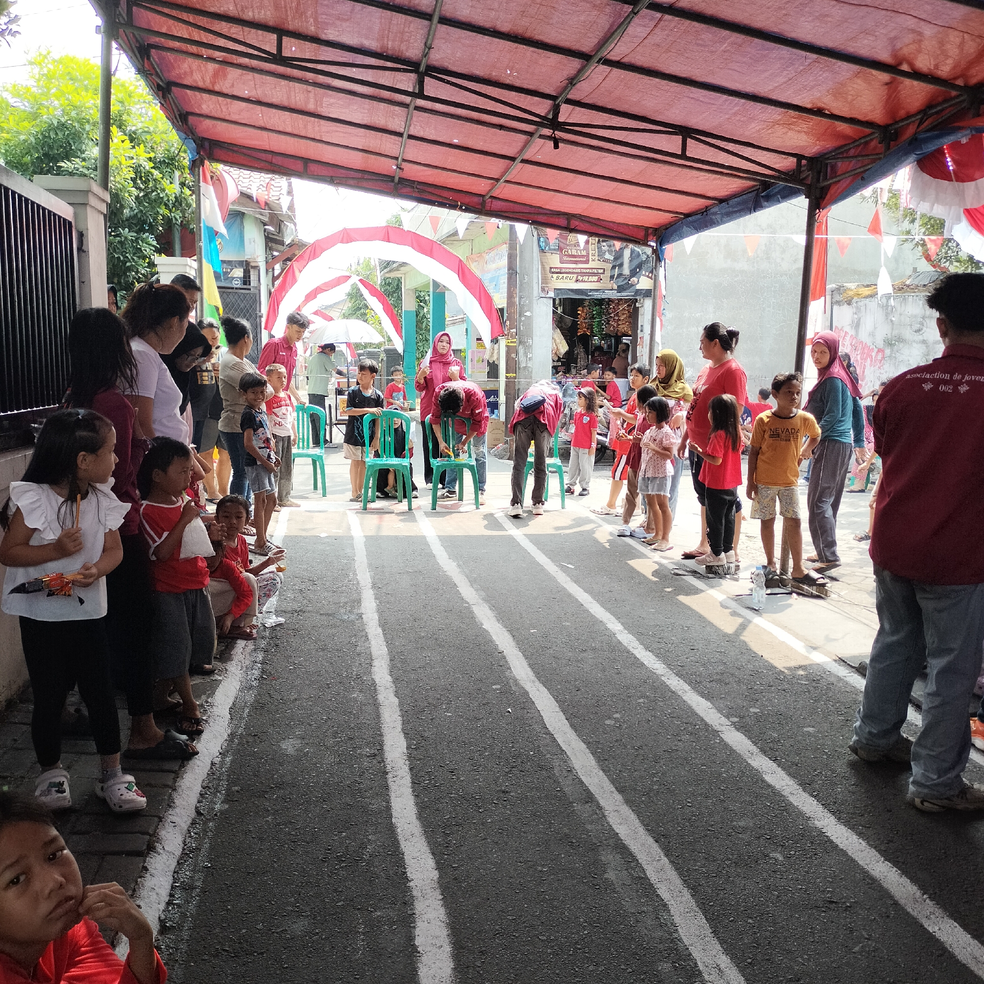 In Tangerang, Indonesia, under a red tarp canopy decorated with Indonesian flags, a group of people are gathered for an outdoor event. The scene appears to be a street or alleyway, with an asphalt road marked with white lines suggesting a racecourse. In the foreground, several children and adults are lined up along the side of the road, with some children participating in a game involving chairs. A boy in a red shirt and black shorts is actively engaged in the game, bent over a green chair. On the right side of the image, a man in a red shirt and jeans stands with his back to the camera, looking towards the event. The environment is bright and sunny, with trees visible in the background, and a building with shops is situated behind the participants. The overall atmosphere suggests a community gathering or celebration, possibly a local competition or festival.