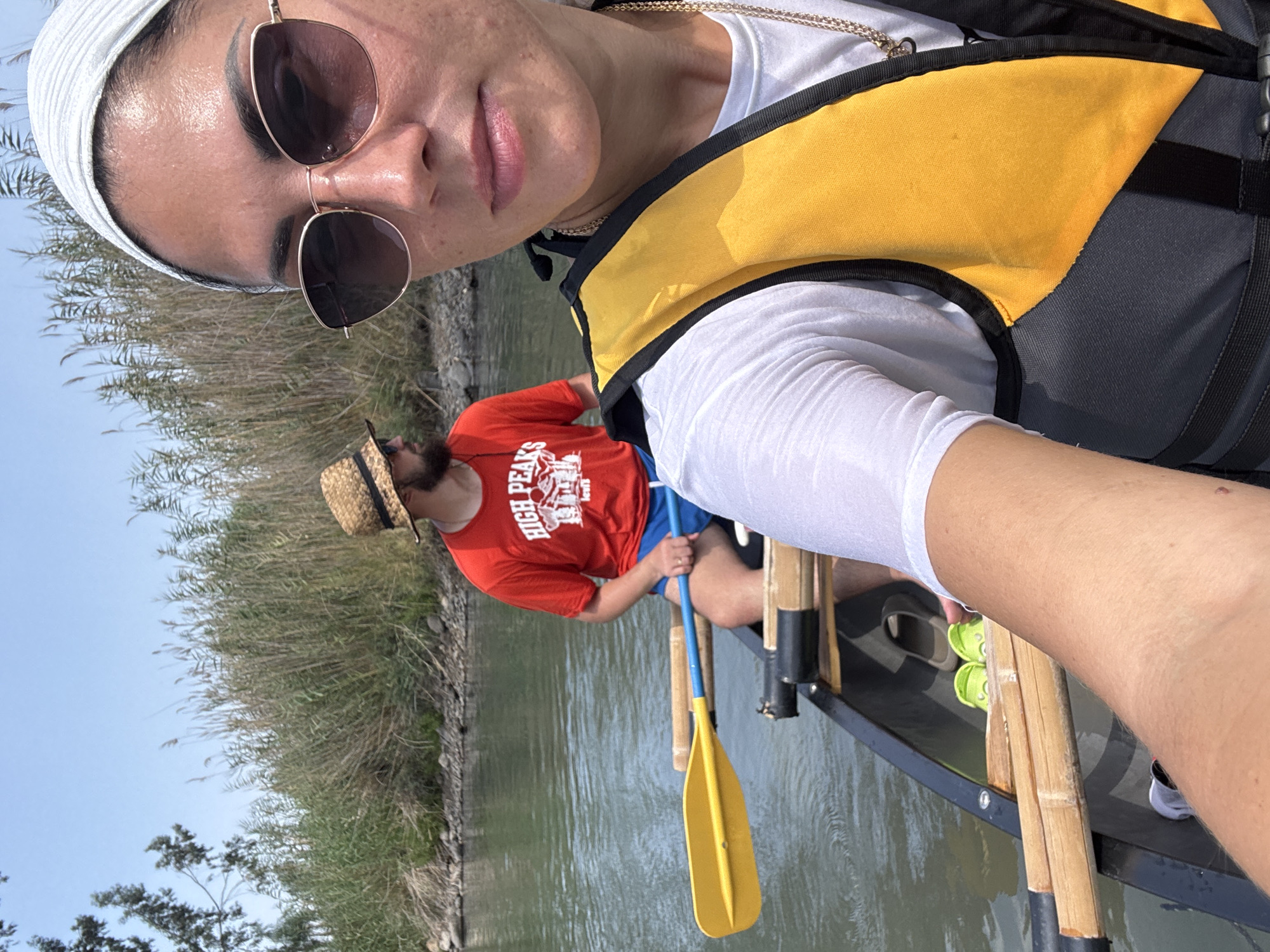 Two individuals are in a canoe on a body of water, possibly the Jordan River in the Emek HaYarden Regional Council, Israel, given the dense reeds and dry, sunny environment. In the foreground, a woman wearing sunglasses, a white headscarf, and a yellow life vest is taking a selfie. Behind her, a man wearing a straw hat and a red t-shirt with "HIGH PEAKS" printed on it is visible. The canoe contains paddles, bamboo poles, and bright green footwear. The water is calm, reflecting the clear blue sky, and the background is dominated by tall, dry reeds and a rocky bank.