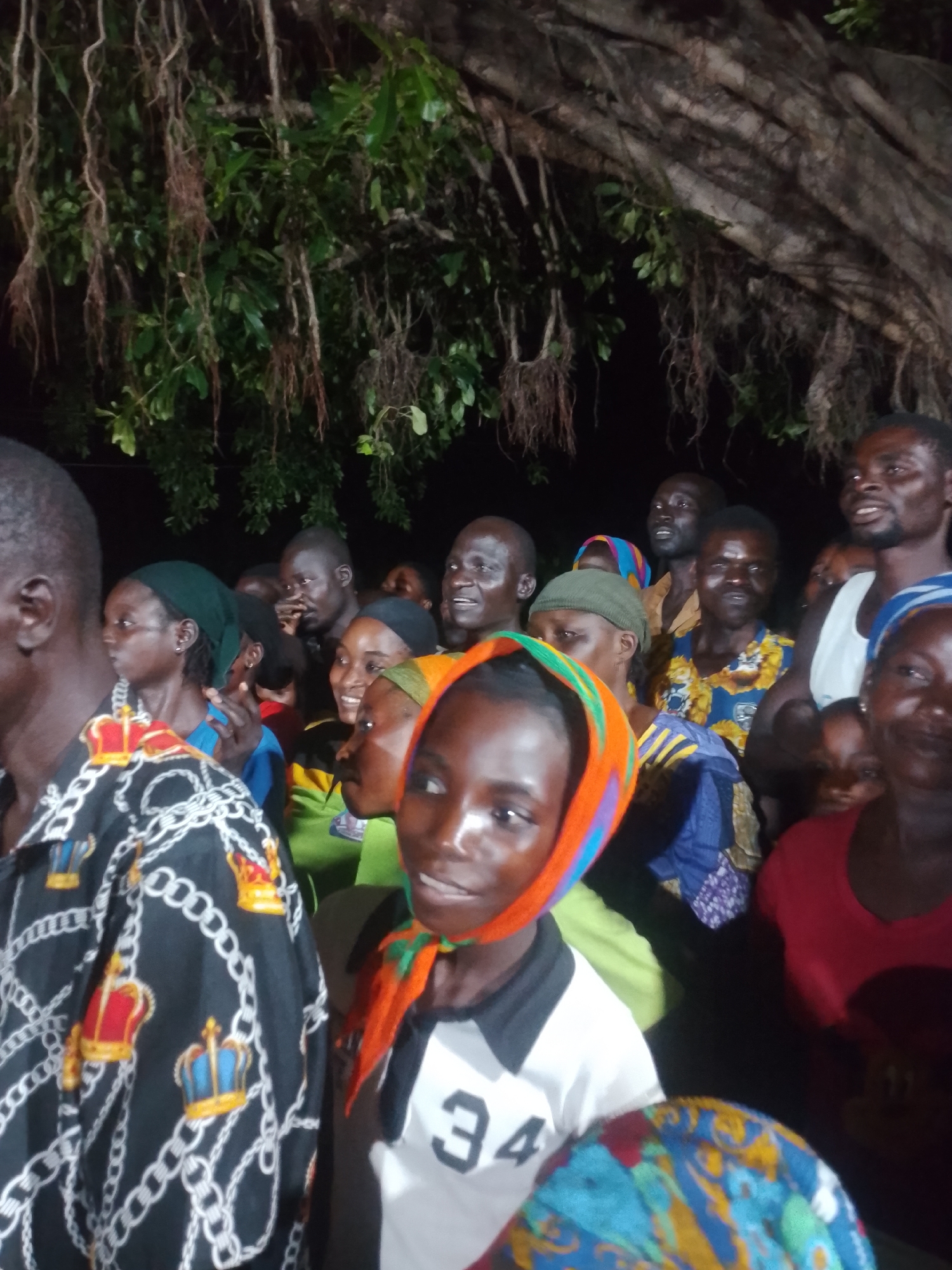 A group of people are gathered outdoors at night, possibly at a public event or meeting. The setting appears to be under a large tree with hanging roots and foliage visible in the upper portion of the image, suggesting a tropical or subtropical environment. The individuals are mostly facing forward, with many looking towards an unseen point or speaker. Some people are smiling and appear engaged in the event. The lighting is artificial and appears to be from a light source off-camera. The clothing styles suggest a casual or traditional attire. The overall atmosphere seems to be one of communal gathering and shared interest.