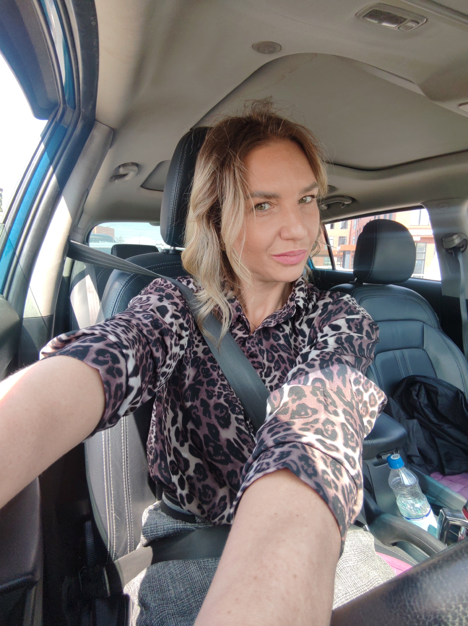 A woman is captured in a selfie taken from the driver's seat of a car during the daytime. She is an adult with blonde or light brown hair, looking directly at the camera with a calm, slightly smiling expression. She is wearing a dark and light brown leopard print long-sleeved shirt and a grey skirt or pants, with a seatbelt fastened across her.
The interior of the car is visible, featuring dark-colored leather seats with light stitching. A sunroof is open above, letting in natural light. Sunlight streams in from the driver's side window, indicating bright, clear weather conditions. On the front passenger seat, a dark bag or jacket is placed. Between the front seats, a plastic water bottle with a blue cap is visible, with partially discernible Cyrillic text on its label, likely indicating "Bonaqua." Some pink fabric is also seen on the console area.
Through the car windows, a background of buildings, trees, and sky suggests an urban or suburban setting, consistent with the location context of Petropavlovsk-Kamchatsky, Russia. The woman is stationary, posing for the camera, possibly before or after a journey. No other individuals are present in the frame. The scene conveys a casual, everyday moment.