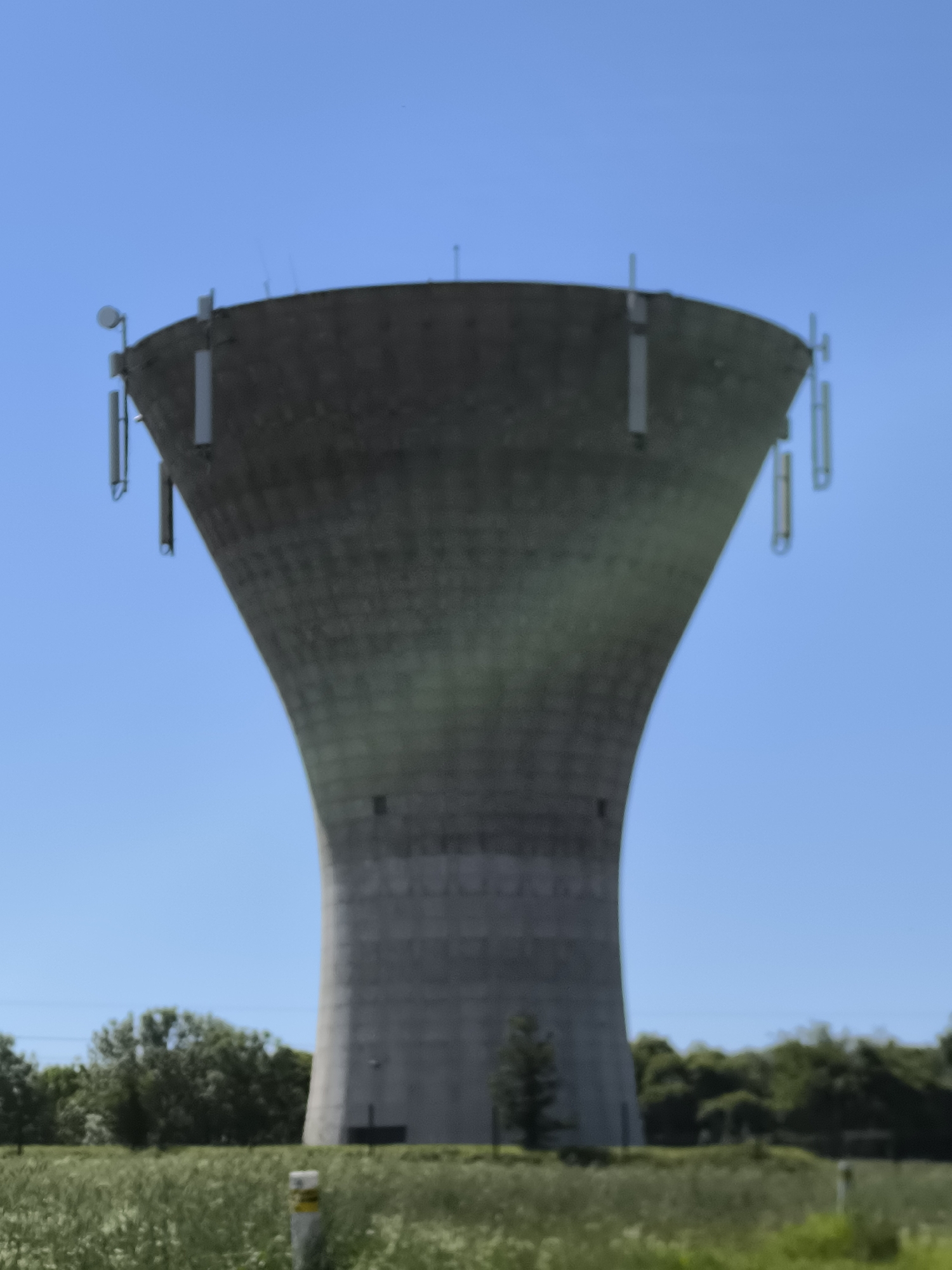 The image shows a water tower in Chaponnay, France. The tower is gray and has a unique shape, wider at the top and narrowing towards the base. Several antennas or communication devices are attached to the top of the tower. The sky is clear blue. In the foreground, there's a field of tall grass and some trees are visible in the background. A small white post or marker is visible near the tower's base.