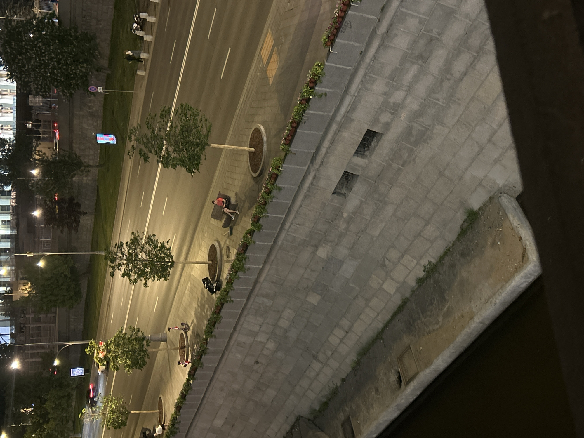 The image shows an elevated view of a city street at night. A road with multiple lanes runs parallel to a pedestrian walkway lined with trees in circular planters. There are two people sitting on a bench. The walkway is paved with gray stone and is bordered by a low wall with vegetation and flowers. A river runs along the right side of the walkway. The image is taken from a high vantage point, looking downward.