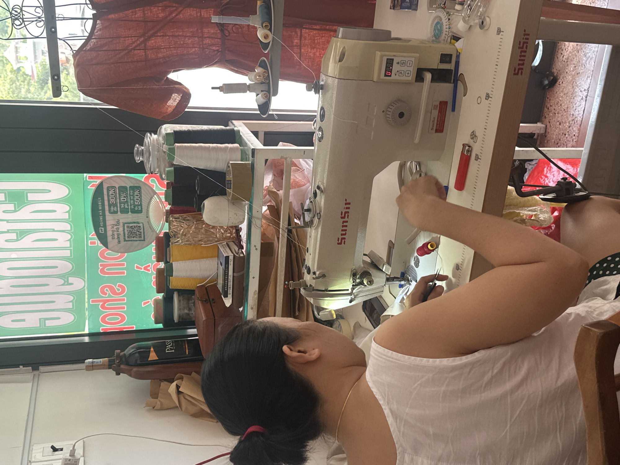 The image depicts a medium shot of a Vietnamese woman in Bac Nha Trang Ward, Vietnam, working at a sewing machine. She has black hair pulled back in a bun and is wearing a white sleeveless top. The sewing machine, a SunSir model, is cream-colored with a digital display. The woman is actively engaged in sewing, holding scissors in her right hand and manipulating fabric with her left.
The environment appears to be a workshop or tailor's studio. On a shelf behind the sewing machine, there are spools of thread in various colors (black, white, yellow, green), a glass jar, and a bottle of wine labeled "PASS." To the right of the sewing machine, a mannequin is dressed in a rusty orange garment. The background includes a large green sign with white lettering, partially obscured. The overall impression is of a functional workspace focused on textile work.