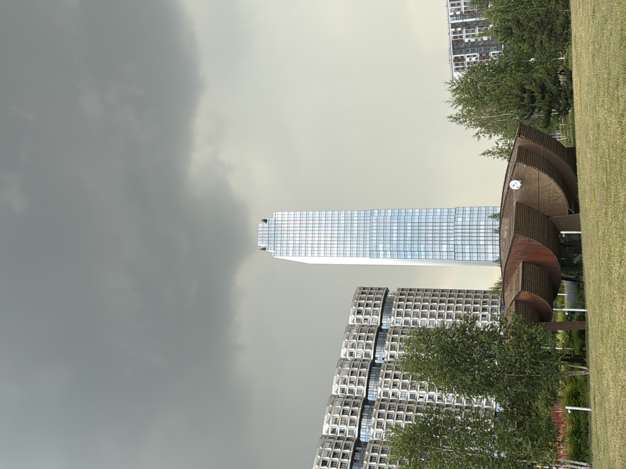 This image depicts a view of the Moscow International Business Center, also known as Moscow City, in Moscow, Russia. In the foreground, there is a park with a grassy lawn and several trees. A distinctive, curved wooden structure, possibly a pavilion or art installation, is situated in the middle ground. Behind the park and the wooden structure, several modern skyscrapers are visible. The tallest is a sleek, glass-faced skyscraper that dominates the center of the frame. To its left, a large, multi-story building with a unique, honeycomb-like facade is prominent. To the right, another residential building with a red and white patterned facade can be seen. The sky is overcast with dark, heavy clouds, suggesting an impending storm. The overall scene captures a blend of urban architecture and green space under a dramatic sky. There are no discernible individuals or specific actions occurring within the frame.