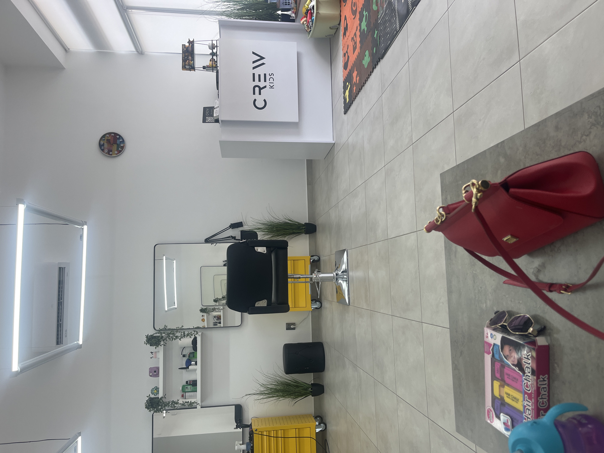 This is an indoor photograph of a children's hair salon. The salon is brightly lit with modern fluorescent lighting. The walls are white, and the floor is tiled with large, light gray square tiles.
In the foreground, on a gray table, is a red handbag with gold hardware. To the left of the handbag is a box of "Hair Chalk" for children, along with a pair of sunglasses. A blue sippy cup is also visible.
The background shows the salon's setup. There are two identical yellow rolling tool cabinets, one on the left and one on the right, each with a mirror above it. Between the cabinets, in the center, is a black salon chair with a chrome base. Next to the chair on the right is another yellow rolling cabinet. Several artificial plants in black pots are placed around the salon.
Further back, a white reception desk with a "CREW KIDS" logo is visible on the right side of the image. A colorful children's play mat is on the floor in front of the desk. A clock with a colorful, cartoonish design is mounted on the wall. An air conditioning unit is installed high on the wall above the mirrors. The overall impression is a clean, modern, and child-friendly environment. There are no people visible in the image. The location is not Uzhhorod, Ukraine, as there are no identifying features to suggest this location.