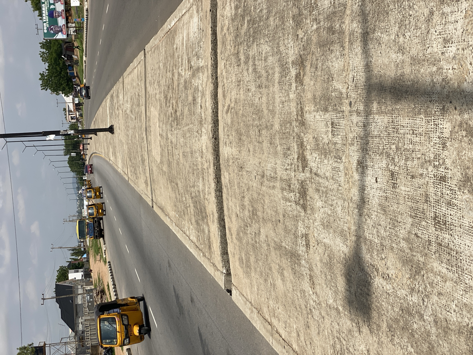 The image shows a street scene in Jalingo, Nigeria, featuring a sidewalk with a weathered concrete surface and a road with several vehicles. In the foreground, the concrete sidewalk shows signs of wear and tear, with distinct joints and an uneven texture. A shadow of a streetlight or overhead structure falls across the sidewalk.
The road, which appears to be a multi-lane highway, is visible to the left of the sidewalk. Several yellow auto-rickshaws, also known as Keke Napep, are traveling on the road, with one prominently positioned closer to the viewer. A yellow truck and other vehicles are also seen in the distance. The road is bordered by a curb and a painted white line.
To the left of the road, there are buildings, some with tiled roofs, and trees. A large billboard with images of political figures and text is visible on the right side of the road in the background. Streetlights line the side of the road. The sky is cloudy, suggesting it might be a warm day. The overall impression is of a busy urban environment in Jalingo.