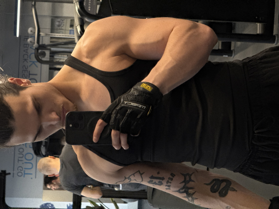 An athletic male, approximately 20-30 years old, with dark long hair tied back, is depicted taking a mirror selfie inside a commercial gym. The gym is identified as likely a "Pulse Fitness & Spa" location in Sofia, Bulgaria, based on the contemporary gym design, dark color scheme, and partially visible motivational wall text such as "GO OUT" and "BACK TO" (seen mirrored in the reflection).
The main subject is positioned centrally, wearing a black ribbed tank top and black athletic shorts with a drawstring. He holds a dark-colored smartphone, likely an iPhone, in his right hand to capture his reflection. His right hand is covered by a black fingerless gym glove with yellow accents, displaying "MECHANIX WEAR" branding. His left arm is visibly muscular and tattooed with the stylized text "EL-13", a thorny vine design, and a coiled snake on the forearm. He looks down at his phone, exhibiting a slightly pouting or focused expression.
In the mirrored background, a second male individual with a shaved head, wearing a grey t-shirt and dark pants, is visible from the back, positioned to the left of the frame. Various pieces of dark-colored gym equipment, including machine frames and weights, are reflected throughout the background. A potted plant is also visible in the far left of the reflection. The floor is a dark gym matting.