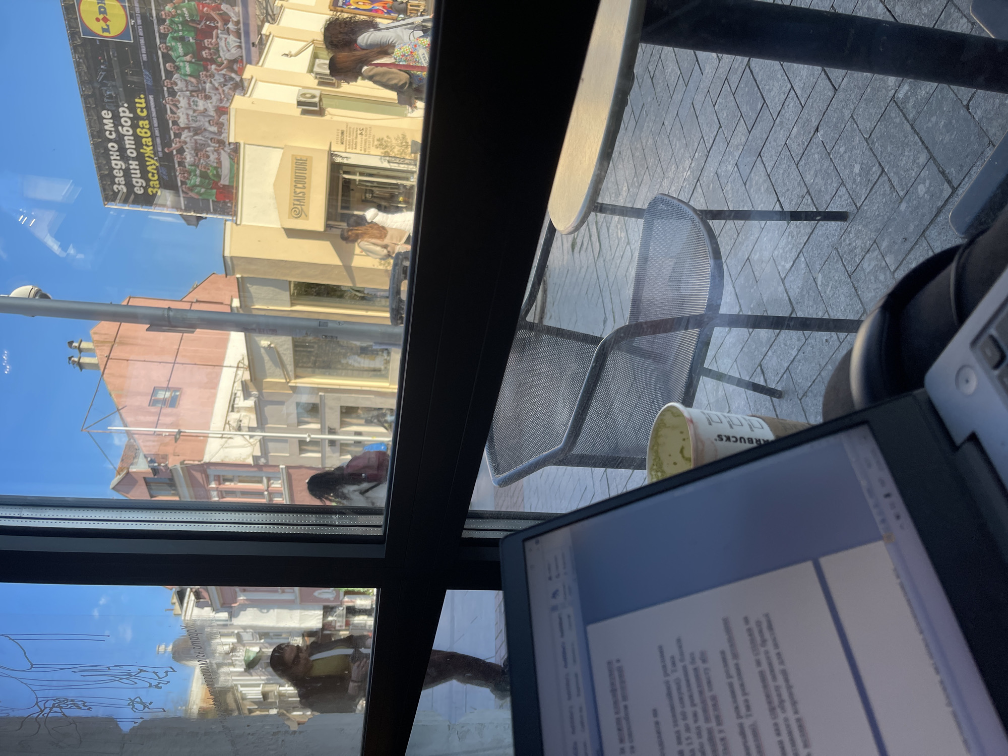 The image captures a vibrant street scene viewed from indoors, looking out through a window onto a sunny urban environment, likely in Varna, Bulgaria. The clear blue sky and bright sunlight indicate daytime with pleasant weather.
Strong reflections on the window foreground reveal elements of the interior, including a person (presumably the photographer) drawing on what appears to be a digital tablet or the window surface itself at the bottom left. A laptop or e-reader screen, partially visible on the right, displays Cyrillic text, alongside a "STARBUCKS" cup and a partial view of headphones, suggesting a work or study setting.
The outdoor scene features buildings with diverse architectures, including yellow and pinkish-brown facades, lining the street. A prominent billboard dominates the upper left, displaying the "LIDL" logo and an image of a sports team in green jerseys. Accompanying Cyrillic text on the billboard reads: "Заедно сме, един отбор. Заслужава си." (Together we are, one team. It's worth it.). Below, storefronts are visible, with signs such as "TAIS COUTURE," and partially obscured "MOSCHINO" and "FURYALL MEN'S WEAR." The street level is paved with grey interlocking bricks in a herringbone pattern, where an outdoor cafe-style table and two metal mesh chairs are positioned. Several people are visible in the distance, walking along the bustling street. The overall impression is a calm, focused indoor activity juxtaposed with the dynamic life of a city street outside.