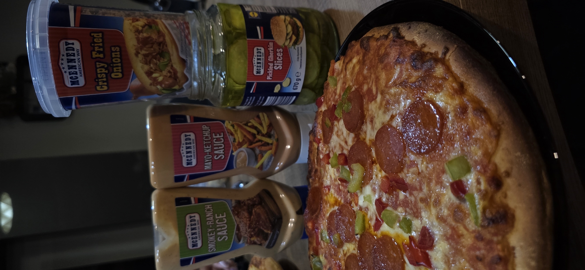 A pepperoni and bell pepper pizza sits on a black plate on a wooden table. To the left of the pizza are two squeeze bottles of sauce: one labeled "Smokey-Ranch Sauce" and the other "Mayo-Ketchup Sauce." To the right of the pizza is a glass jar of "Pickled Gherkin Slices" and a blue and red tub of "Crispy Fried Onions." The background is dark and out of focus, with a ceiling light fixture visible at the top. This image was taken indoors, likely in a home kitchen or dining area, and does not contain any information about its location in Vratsa, Bulgaria.