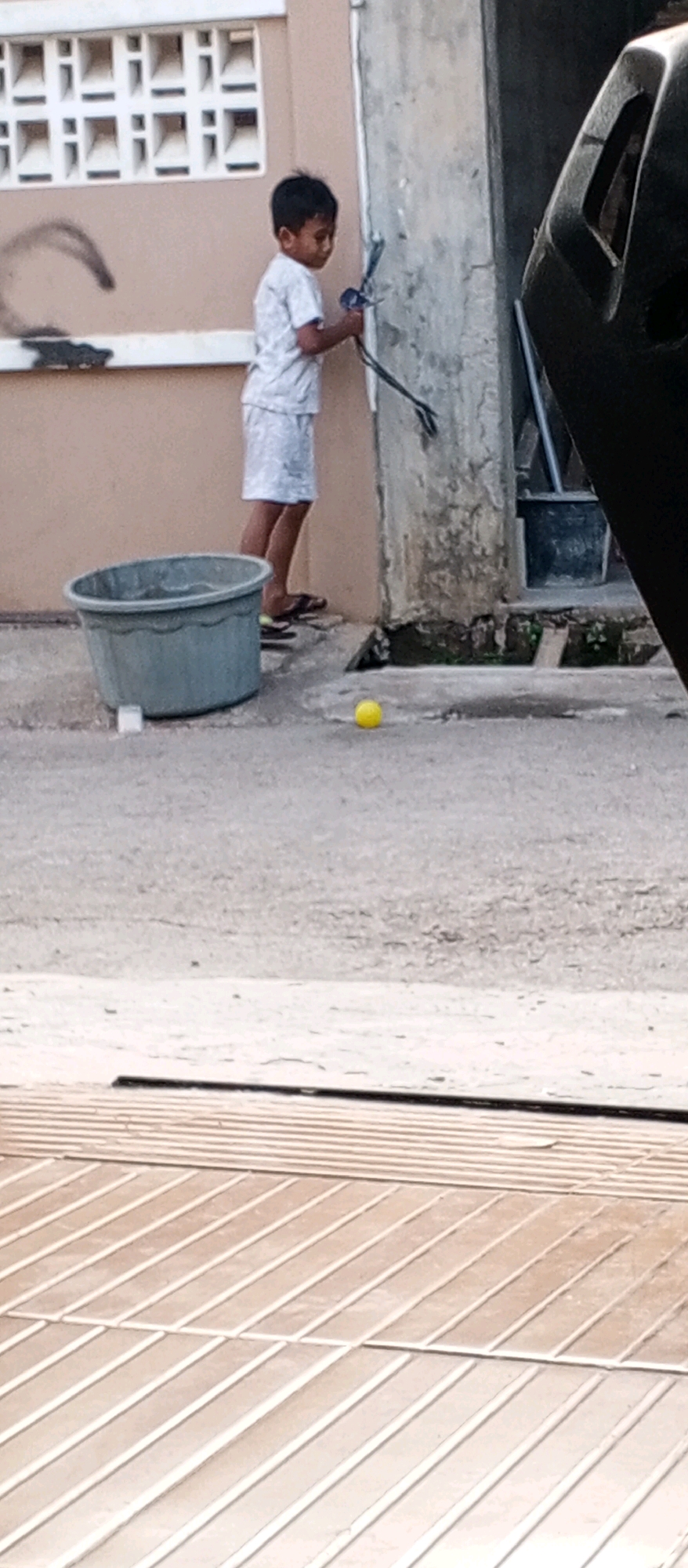 A young boy in a white t-shirt and shorts stands on a concrete surface, holding a toy bow and arrow. To his left is a large, grey plastic tub. A yellow ball rests on the ground in front of him. Behind him, a light brown building with a patterned window is visible. To the right of the boy, a black object, possibly part of a vehicle, is partially visible. The scene appears to be outdoors, likely in a residential or urban area, with a focus on the boy and his toy.