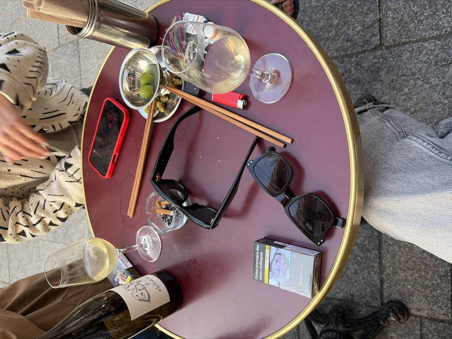 The scene captures a close-up, downward-angled view of a dark red, round outdoor cafe table with a golden rim, located in Paris, France.
Arranged on the table are multiple items. On the left, a green-tinted wine bottle, labeled "B... à la fraîche", stands next to a wine glass containing white wine. Nearby rests an open pack of Marlboro Red cigarettes displaying the French health warning "Fumer provoque des crises cardiaques". A glass ashtray holds several cigarette butts. Two pairs of dark sunglasses are present: one folded, with a visible Ray-Ban style, and another, labeled "KALEOS", lying open. Two sets of wooden chopsticks are positioned centrally. A red-cased smartphone displays the time 18:27 on its dark screen. To the right, a small metal bowl on a saucer contains two green olives and several olive pits. Adjacent to this, a tall stainless steel cylinder holds rolled paper items, possibly napkins. Another wine glass, partially filled with white wine, a red cigarette lighter, and small tubes labeled "Aqua..." are also visible.
Partial views of individuals are present: a person wearing a brown garment is visible on the far left, and a person in a white and black geometric patterned jacket with manicured hands is visible on the opposite side of the table. The legs of the photographer, clad in light gray jeans, are visible beneath the table, resting on gray pavement.