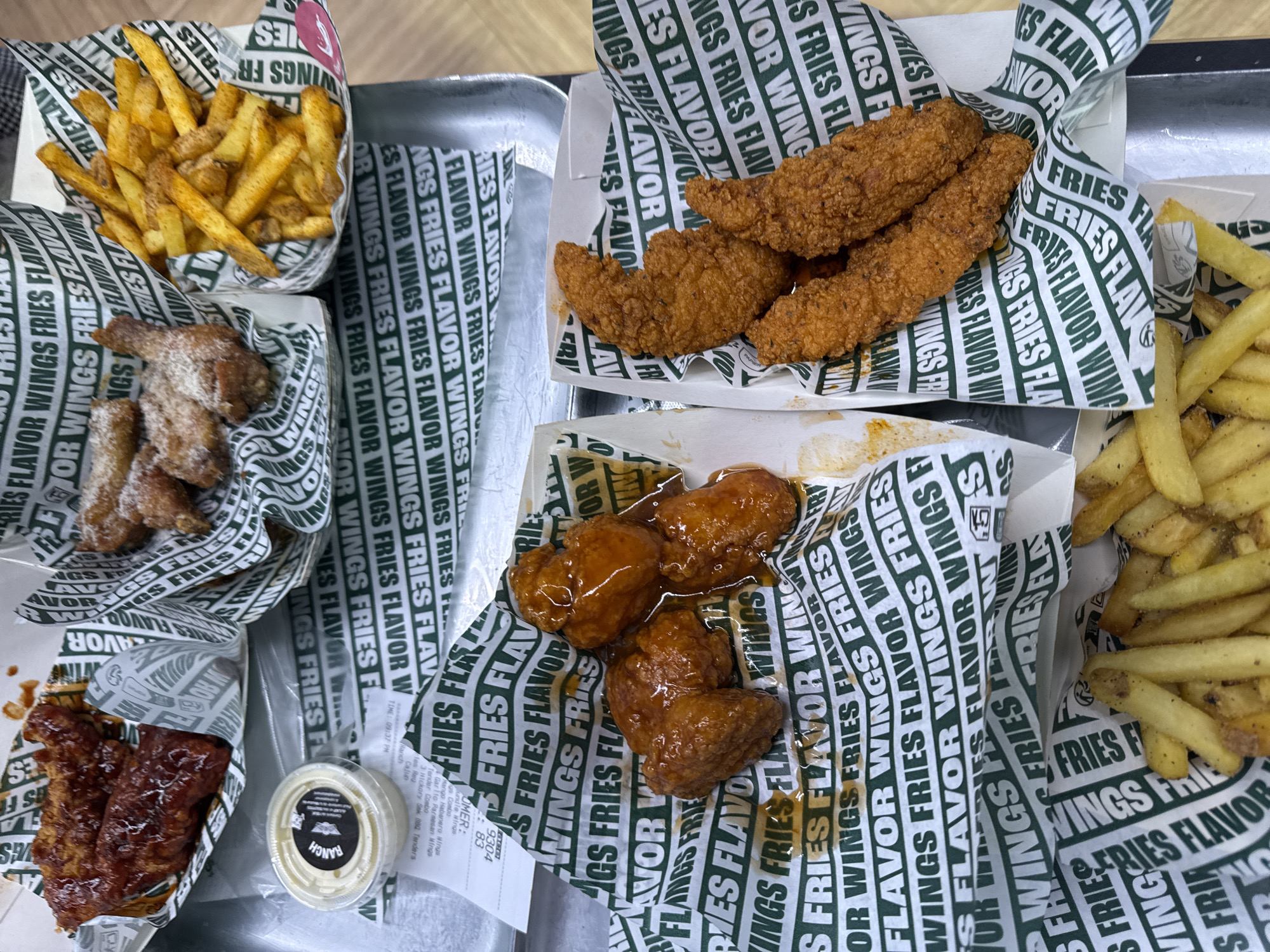 The image shows a metal serving tray covered in paper liners printed with "WINGS FRIES FLAVOR WINGS FRIES FLAVOR". On the tray are multiple containers of food, including:
* One container filled with french fries.
* One container holding wings covered in a light-colored, powdered coating.
* One container filled with what appear to be barbecue-glazed wings.
* One container holding chicken tenders.
* One container filled with what appear to be honey-glazed wings.
* Another container filled with french fries.
* A small container of Ranch dipping sauce.
* A receipt.
There is no indication of location within the city of Paris, France.
