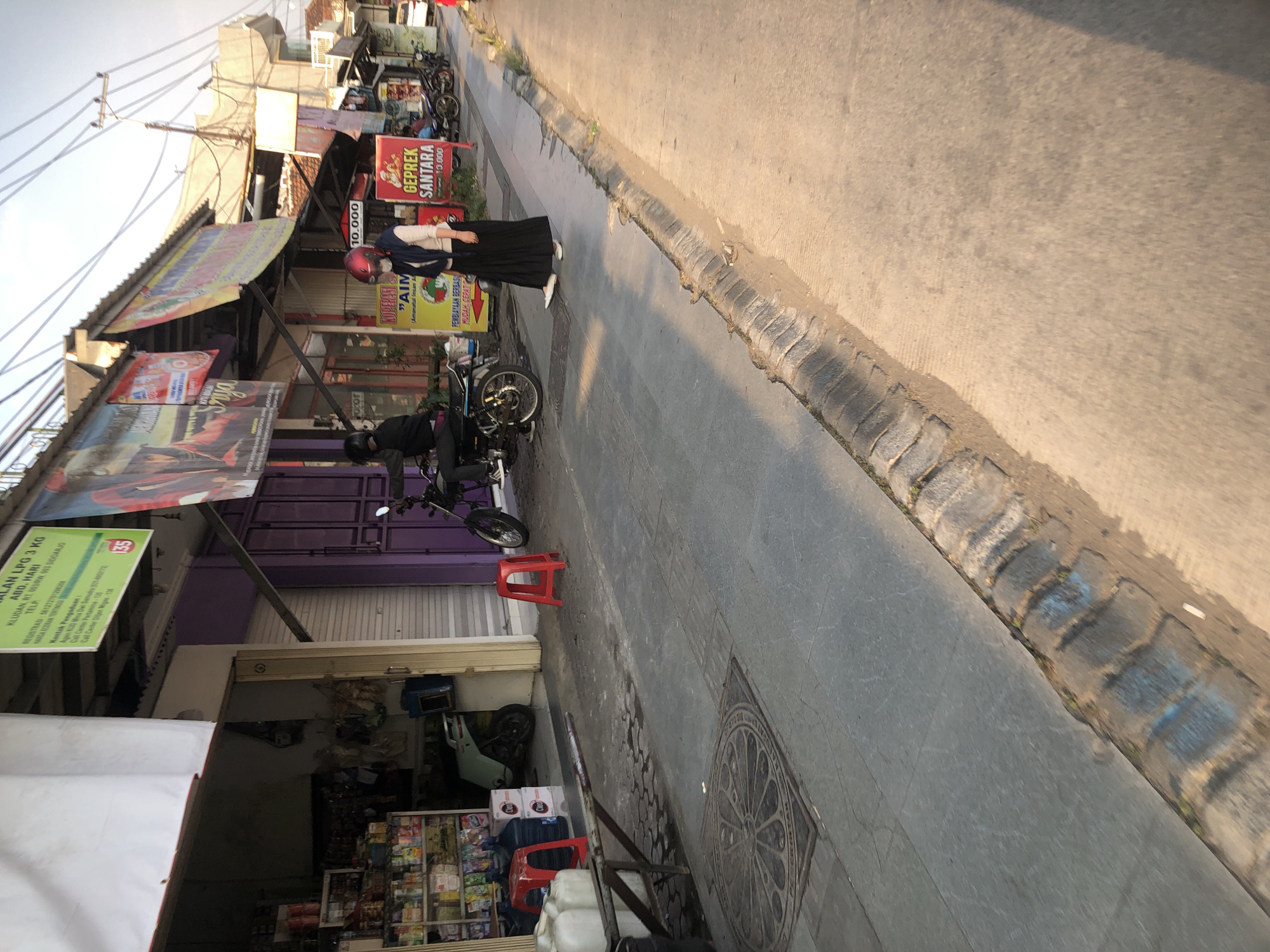 The image shows a street scene in Boro, Indonesia, with several small shops lining the left side of the frame. In the foreground, a gray paved sidewalk is visible, with a circular metal grate in the pavement. Red plastic stools are placed on the sidewalk. A person wearing a black jacket and a helmet is seated on a motorcycle parked in front of one of the shops. Further down the street, another person wearing a white long-sleeved shirt, a dark headscarf, and a long dark skirt is walking on the sidewalk. They are wearing white shoes and a face mask. Several shops have colorful banners and signs displayed. One banner shows text in Indonesian and an image of a woman. Another sign advertises "GEPREK SANTARA" with prices in Indonesian Rupiah. A motorcycle is parked in front of this shop. The street is paved with asphalt, and there is a concrete curb separating the sidewalk from the road. The sun is casting long shadows, suggesting it is late afternoon or early morning. The overall environment appears to be a commercial area with modest buildings and a somewhat busy street.