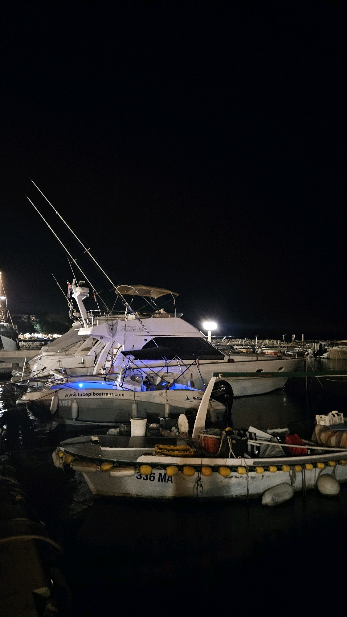 At night, in the marina of Općina Tučepi, Croatia, several boats are docked. The largest is a white yacht with a tan canopy, featuring fishing rods and a blue light under its deck. In front of it, a smaller white boat with a dark hull is also illuminated by blue lights, displaying the website "www.tucepiboatrent.com" and the marking "MA". Further in the foreground, a weathered, gray boat with yellow buoys along its side is visible, marked with "336 MA". The background shows distant lights of the town and other docked boats. The water is dark and still, reflecting the lights from the vessels. There are no people visible in the image.