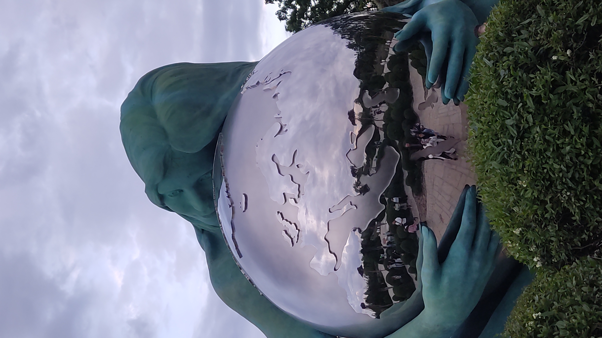The image displays a monumental teal-colored statue, likely a woman or goddess, holding a large, highly reflective spherical globe. The statue's serene face is visible in the upper left, gazing downward at the globe cradled in its hands. The teal material of the statue appears weathered, suggesting a patinated bronze or similar finish.
The polished surface of the globe acts as a mirror, vividly reflecting its surroundings. Etched outlines of continents, including Europe, are visible on its surface, indicating it represents the Earth. Within the reflections, a paved area with several pedestrians, trees, and a cloudy sky are distinctly visible, offering a clear view of the park environment surrounding the sculpture. A dense, dark green bush with small white flowers occupies the right side of the frame, providing a natural contrast.
The sky is overcast with soft, diffuse clouds, indicating daylight conditions. The perspective is tilted, creating a dynamic composition of the figure seemingly holding the entire world. The sculpture, known as "Mother Earth" or "Gaia," is located in a public park in Krasnodar, Russia, and conveys a sense of guardianship and reverence for the planet. No visible text is present in the image.