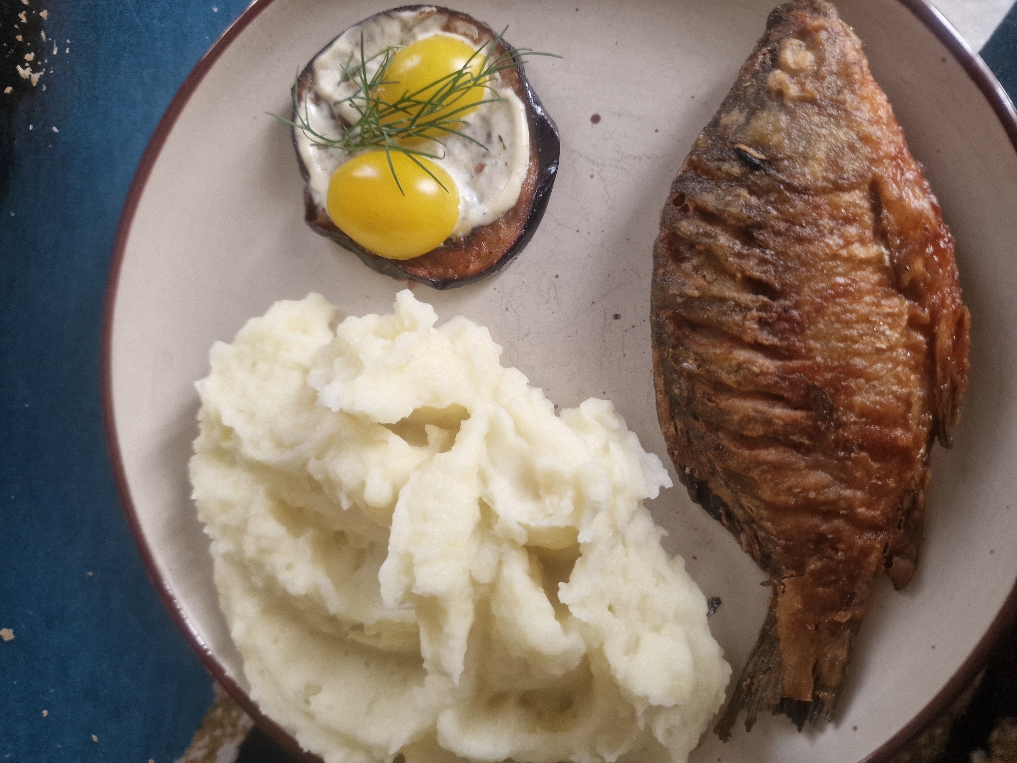 A close-up overhead shot shows a plate of food. The plate is light-colored with a dark rim and rests on a dark blue surface with specks of gold. In the upper left of the plate is a mound of mashed potatoes. To the right of the potatoes is a slice of grilled eggplant topped with a creamy sauce, two small yellow cherry tomatoes, and a sprig of dill. In the lower portion of the plate is a whole fried fish, positioned horizontally. The fish appears golden brown and crispy. The overall scene is a top-down view of a prepared meal. There are no identifiable landmarks or features to indicate the city of Makinsk, Kazakhstan, or any specific urban location.