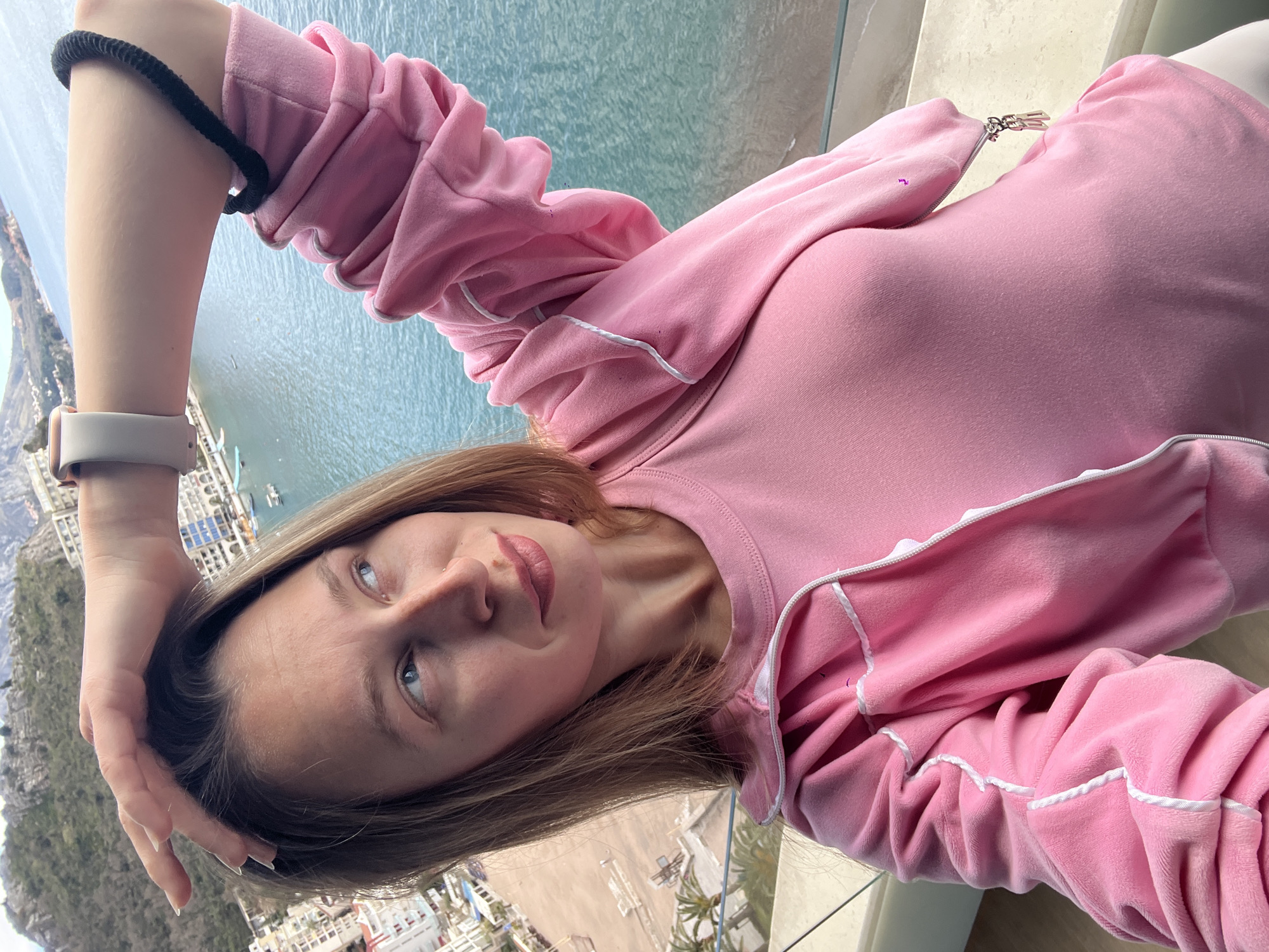 A woman is photographed from a high angle, likely on a balcony overlooking a coastal scene. She is wearing a pink velour tracksuit with white piping. She has blonde hair and is wearing a light-colored watch on her left wrist. Her left arm is raised, with her hand near her forehead. The background shows a town by the sea, likely Kamenovo, Montenegro, judging by the coastal landscape and building styles. The sea appears calm, with a few boats visible. Part of a clear balcony railing is visible. The scene suggests a relaxed, tourist setting.