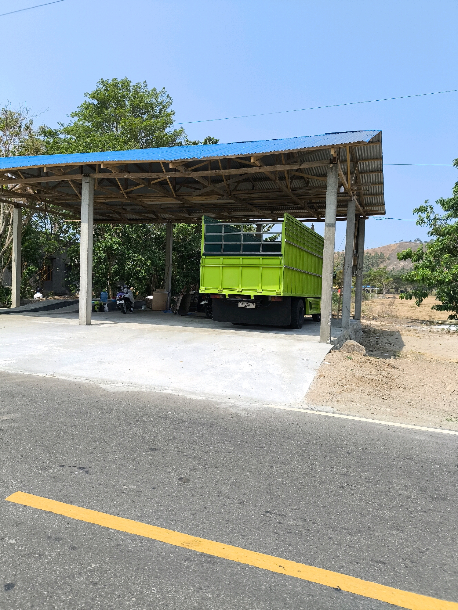 A bright green truck with a high-sided cargo bed is parked under a covered structure with a blue corrugated metal roof. The structure is supported by concrete pillars and wooden beams. In the background, there are trees and a dry, hilly landscape. A yellow line is painted on the asphalt road in the foreground. A motorcycle is parked to the left of the truck under the shelter. The sky is clear and blue.