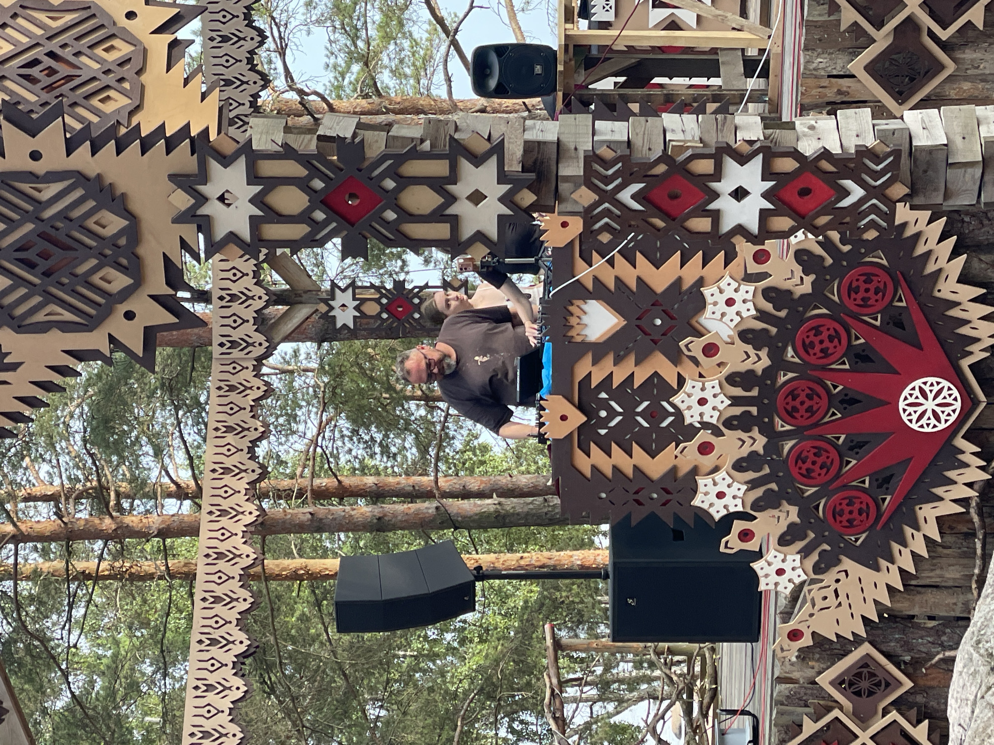 This image depicts an outdoor DJ performance at an event in Luzhki, Russia, characterized by a rustic and nature-inspired aesthetic. In the foreground, a DJ, a man with glasses and a beard, is actively mixing music at a console. He is wearing a dark t-shirt. Beside him, a young woman with light brown hair stands looking towards the DJ. They are positioned behind a DJ booth decorated with intricate, laser-cut wooden panels featuring geometric patterns in brown, red, and white. Two large black speakers are visible, one on a stand to the left of the DJ and another smaller speaker on a table to the right. The background is dominated by tall pine trees, suggesting a forest or park setting. Further decor includes more laser-cut wooden elements with symbolic patterns, reminiscent of traditional folk art, suspended above and integrated into the stage structure. The overall scene conveys a festive, open-air atmosphere.