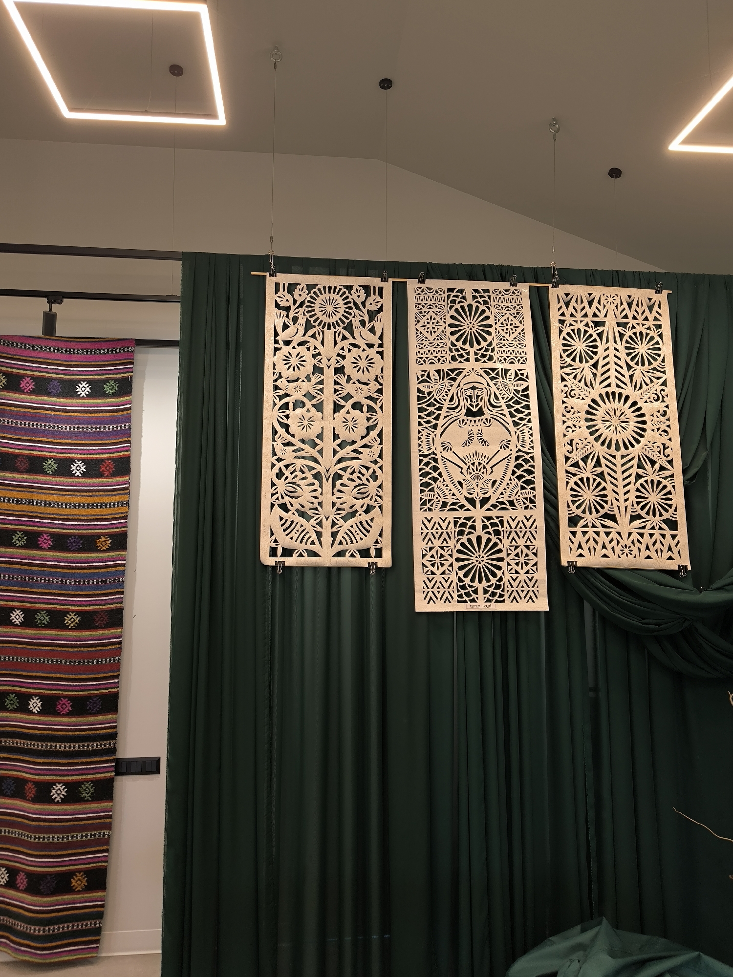 The image shows a display in an indoor setting, featuring traditional Ukrainian art. On the left, a colorful, striped woven textile hangs vertically. To the right, three light brown, intricately cut wooden panels hang suspended in front of a backdrop of dark green, draped fabric. The center panel depicts a woman in a traditional folk style. All three panels are held in place by clips. The setting is illuminated by overhead square LED lights. Based on the text visible on the center panel, it is likely located in Vinnytsia, Ukraine.