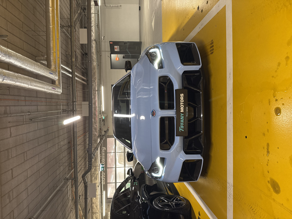 An indoor scene depicts a light grey/blue (BMW Brooklyn Grey or similar) 2023+ BMW M2 (G87) coupe parked head-on in a designated parking bay. The vehicle's headlights are illuminated. Its prominent front grille features a rectangular design, and the lower bumper integrates black carbon fiber elements with large air intakes. The BMW emblem on the hood includes a surrounding M-color ring (light blue, dark blue, red). A black plate holder centered on the grille displays "TIFFANY MOTORS" in white text.
To the left, the front quarter of a dark-colored sedan, equipped with black multi-spoke wheels, is partially visible. In the background, behind the M2, a glass garage door is visible, above which text reading "auto & detailing" is illuminated. To the right, a dark grey door is marked with a red triangular sign and the Polish word "BIURO" (Office) on the wall above it.
The facility features a concrete ceiling with exposed silver-insulated ventilation ducts, various pipes, and linear fluorescent light fixtures providing artificial illumination. The floor is painted bright yellow with crisp white lines demarcating the parking space. Several dark, irregular liquid stains are present on the yellow floor directly in front of the BMW.
Based on the "TIFFANY MOTORS" branding and the "BIURO" sign, the location is a car detailing or showroom facility associated with Tiffany Motors, a luxury car dealership specializing in BMW M vehicles in Warsaw, Poland.