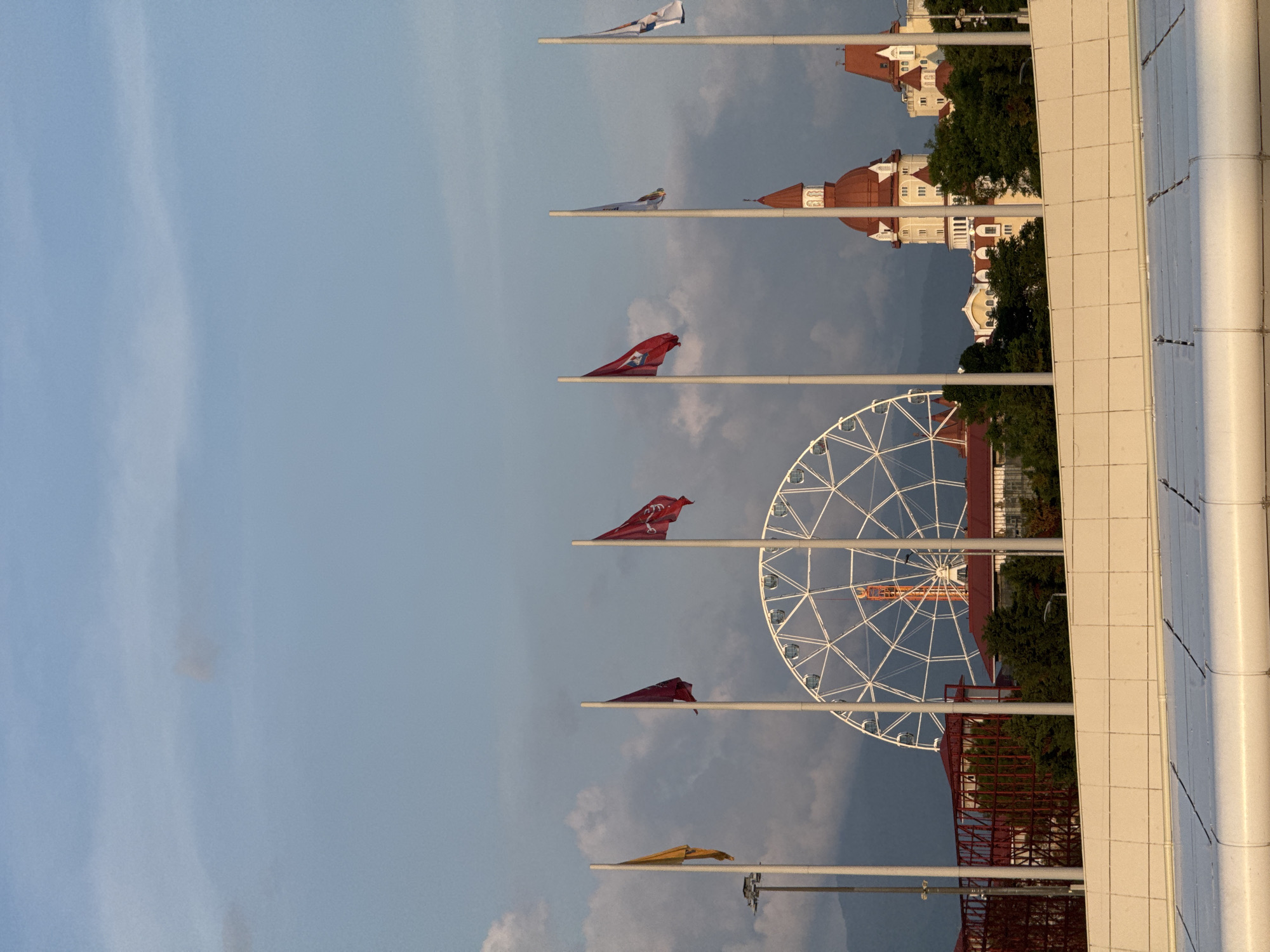 In the city of Sirius, Russia, a Ferris wheel stands tall against a cloudy sky. Several flags on poles are visible in the foreground. In the background, two buildings with red roofs and ornate architecture rise above a line of trees. The scene appears to be a public space, possibly a park or amusement area.