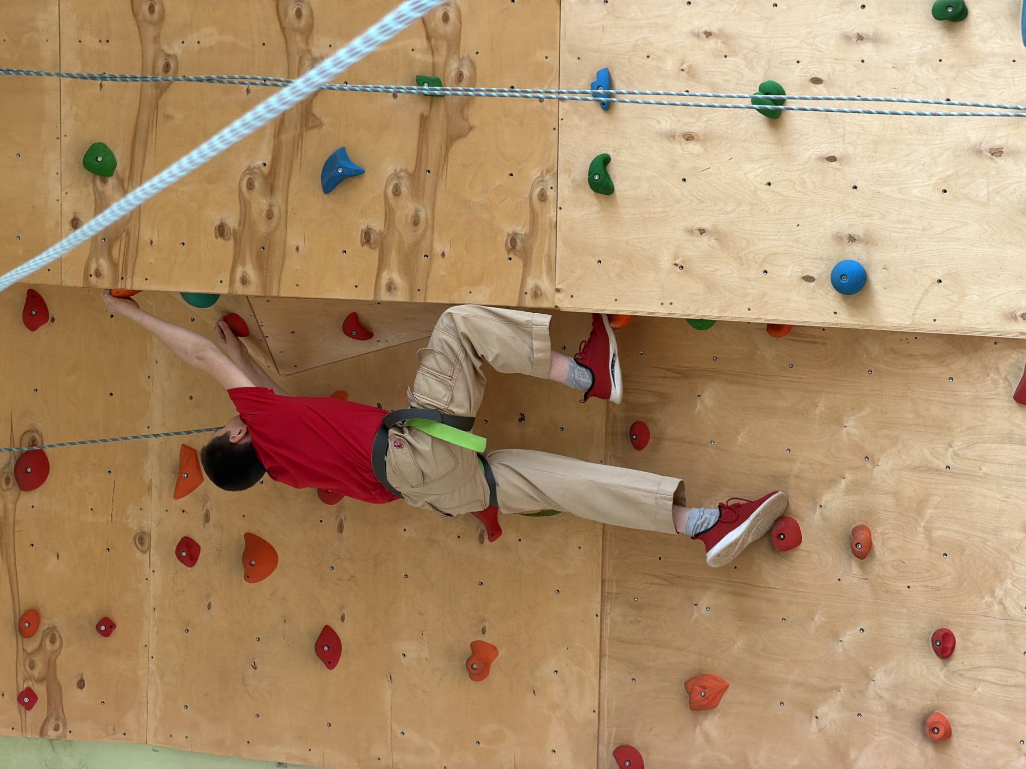 A person is shown climbing on a wooden climbing wall, with various colored holds attached. The climber is wearing a red t-shirt, khaki pants, and red shoes. They are using a harness and rope. The climbing wall is made of light-colored wood panels. The location is not identified, nor is it possible to determine if it is in Izhevsk, Russia.