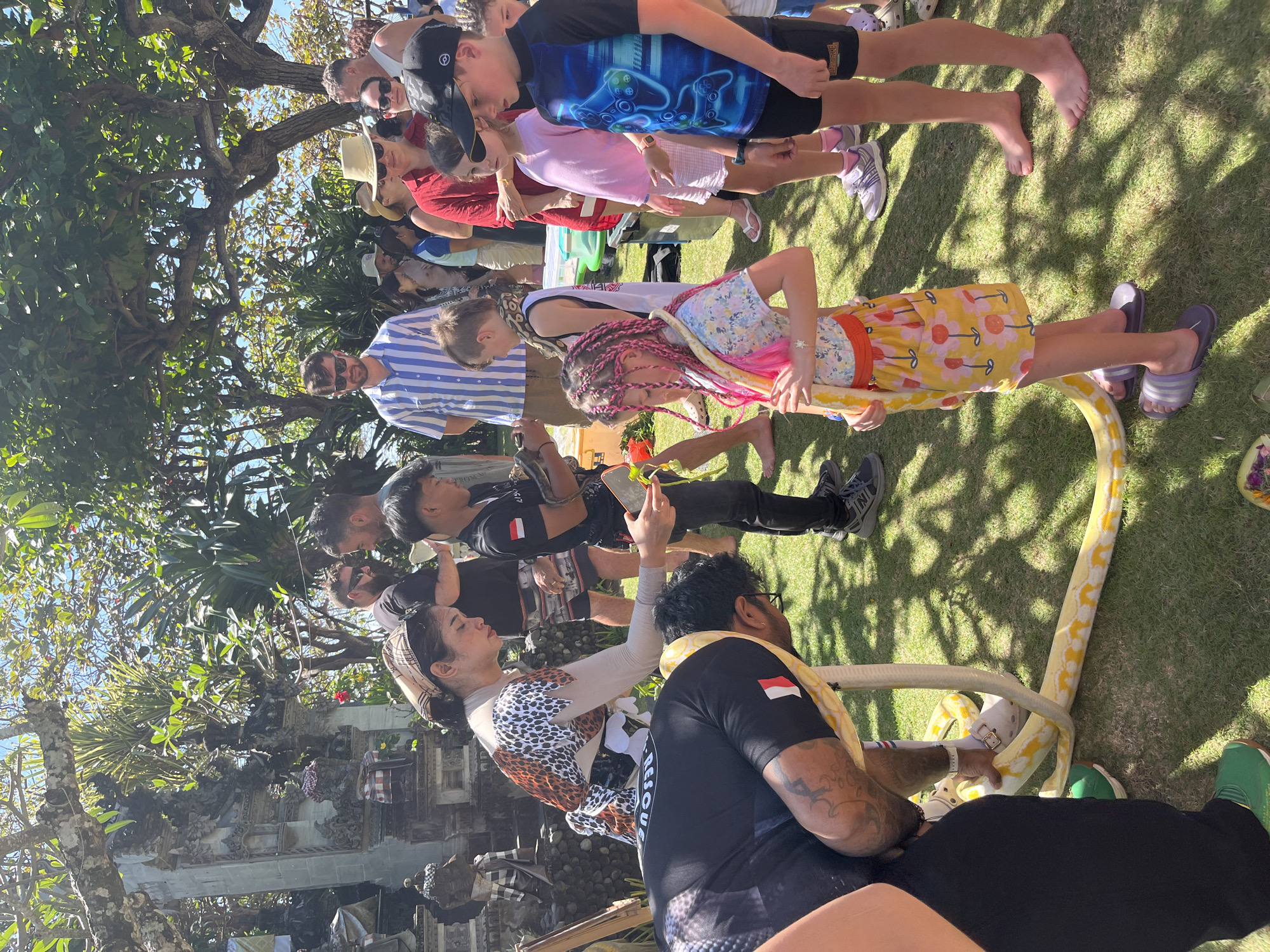In Kuta Selatan, Indonesia, under a canopy of dappled sunlight filtering through lush trees, a group of people, both adults and children, are gathered on a grassy lawn. A large yellow python is the central focus of the scene, being handled by a person wearing a black shirt with white and red markings. Another person, dressed in a leopard-print top and a headscarf, appears to be guiding or assisting with the snake. Several children are watching the snake, some with visible interest and others with more apprehension. The background features elements of traditional Balinese architecture, including carved stone structures and intricate stone work, suggesting a temple or cultural site. The atmosphere appears to be one of a tourist attraction or educational demonstration, with people dressed in casual attire, including shorts, t-shirts, and hats, appropriate for the warm, tropical climate.