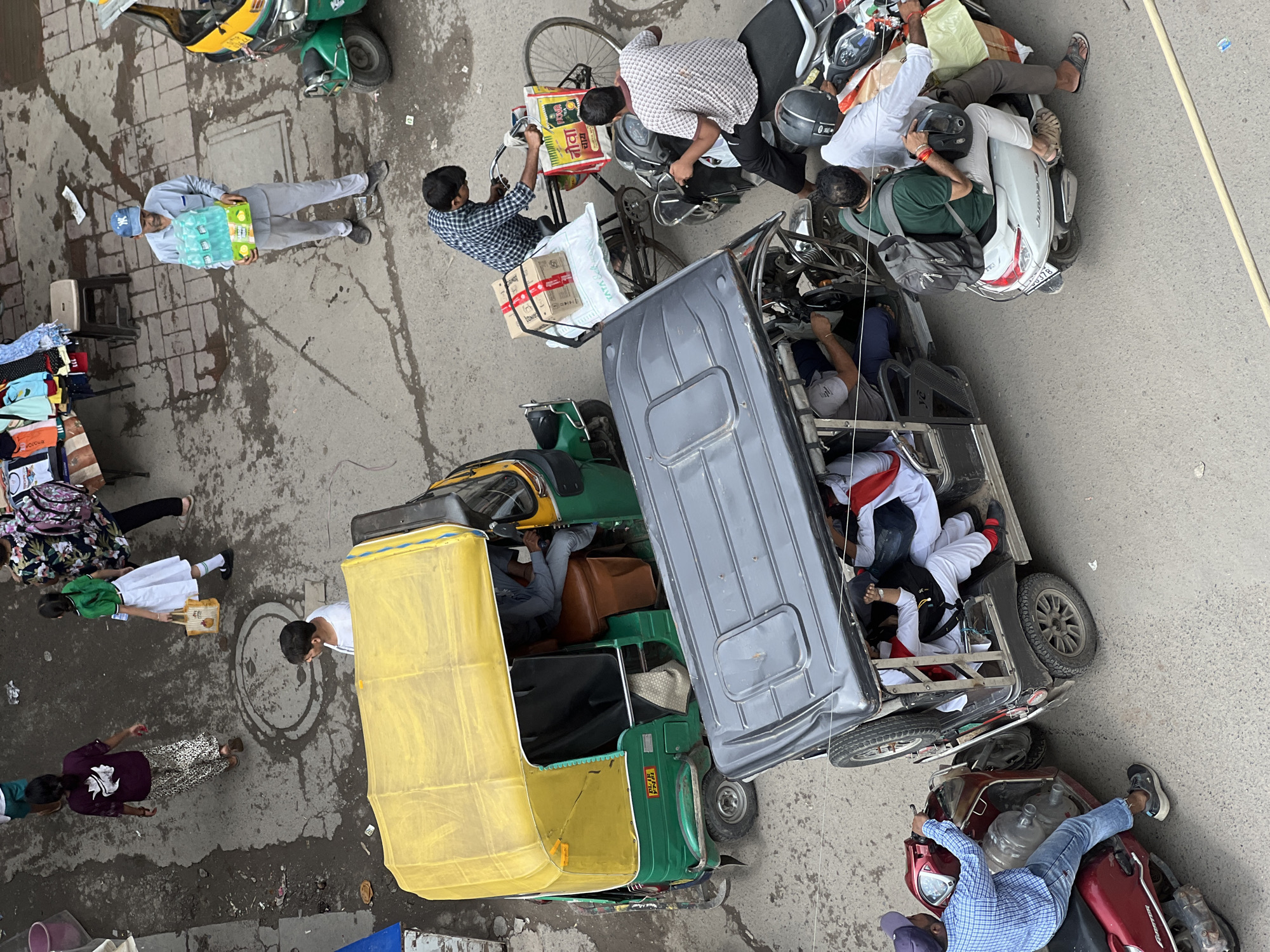 This image captures a bustling street scene in Delhi, India, viewed from an elevated perspective. The foreground is dominated by a dark-colored auto-rickshaw, its roof creating a shadow over the seated passengers. To the left, a red scooter is parked, with a person in a blue checkered shirt and jeans seated on it, holding onto the handlebars. To the right of the auto-rickshaw, a white scooter carries two passengers. The front passenger, wearing a white shirt, is looking forward, while the person behind, in a green shirt and light-colored pants, has a backpack and is holding a helmet. Further back, a green auto-rickshaw with a yellow roof is parked. A man in a blue shirt and cap is walking away from it, carrying a multi-pack of water bottles. A person wearing a green shirt and white skirt is walking with a yellow bag. A woman in a purple shirt and patterned pants is also walking. Behind them, a street vendor's stall displays various colorful clothing items. The ground is a rough, paved surface, with visible tire tracks and some scattered debris, indicating a typical urban street environment. The overall impression is one of daily life in a busy Indian city, with various modes of transportation and people going about their business.