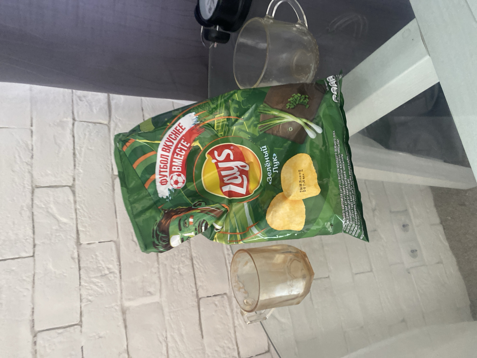 A bag of Lay's green onion flavored potato chips is placed on a glass table in front of a white brick wall. Two empty glass cups are on the table, one to the left of the chip bag and one to the right. The chip bag features a graphic of a person with green and white face paint, and the text "ФУТБОЛ ВКУСНЕЕ ВМЕСТЕ" (Football tastes better together). The overall setting suggests a casual indoor environment. There is no visible indication of the specific location within the city of Saratov, Russia.