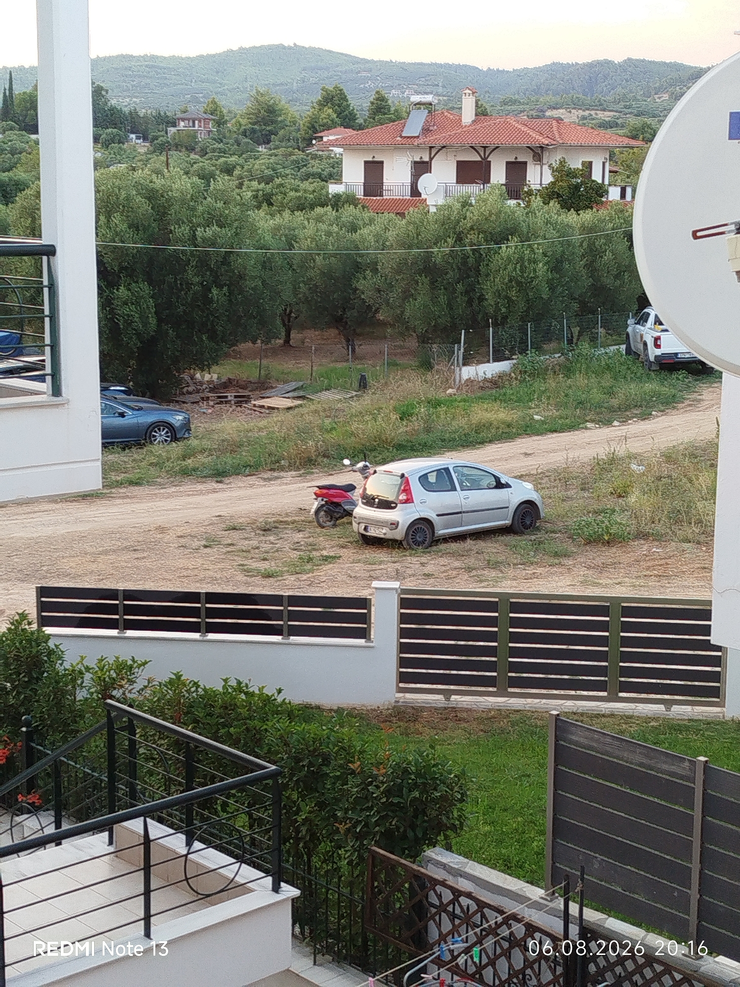 The image depicts a residential area in the Sithonia Municipal Unit, Greece. In the foreground, there is a white building with a balcony railing. Beyond the railing, a dirt road is visible, with a silver hatchback car parked on the right side of the road. A red and white scooter is parked behind the silver car. Further back, a white pickup truck is parked on a dirt track. In the middle ground, a dense row of olive trees and other foliage obscures parts of the scene. In the background, a white house with a red tile roof is situated on a hillside, surrounded by trees. The sky is overcast. The image also contains a large white satellite dish on the right side. The bottom left of the image has a watermark that reads "REDMI Note 13" and the bottom right has a watermark that reads "06.08.2026 20:16".
