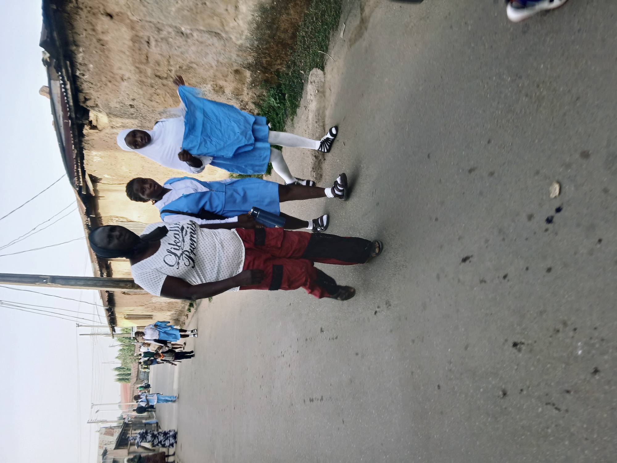 The image, originally rotated 90 degrees counter-clockwise, shows an outdoor scene on a street or pathway in what appears to be a community. In the foreground, three individuals are present. A man stands prominently on the left, wearing a white t-shirt with "Always Promise" printed on it in a stylized font, and red and black pants with black patches, along with dark sandals. To his right and slightly behind him are two young girls, both dressed in blue and white school uniforms.
The girl closer to the man wears a blue uniform top over a white blouse, dark trousers, and black and white sandals. She holds a blue water bottle or flask. The other girl wears a white head covering (hijab) and white leggings under her blue uniform dress, with similar black and white footwear. She is holding up a piece of blue fabric, possibly a scarf, and has a playful expression. Both girls have dark skin and hair.
The ground is a gray, somewhat uneven road surface with various small dark marks. To the right of the subjects, a patch of green grass and dirt is visible. The background features simple residential buildings with textured, light brown walls and some corrugated iron roofs, suggesting a less developed setting. Utility poles and overhead wires line the street. Further down the road, several other people, including children in school uniforms, can be seen, indicating a lively community environment. The natural light suggests it is daytime, possibly under an overcast sky, as shadows are soft or absent. The scene captures a moment of daily life, potentially involving children on their way to or from school.