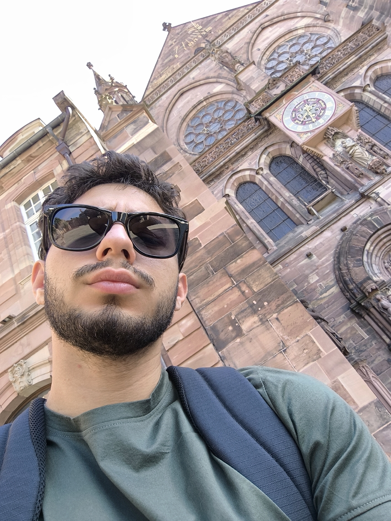 This is a selfie taken from a low angle, with the top of a man's head and shoulders in the foreground. He is wearing a green t-shirt and a dark backpack. He has dark hair, a beard, and is wearing black sunglasses. Behind him is the facade of a large, ornate stone building, likely a cathedral or church, with arched windows and intricate architectural details. A clock face is visible on the building's facade. The stone of the building is a reddish-brown color. The sky above is bright white, suggesting it is daytime. The scene appears to be outdoors in a European city, specifically Strasbourg, France, given the architectural style. The man's expression, as visible through his sunglasses, is neutral.