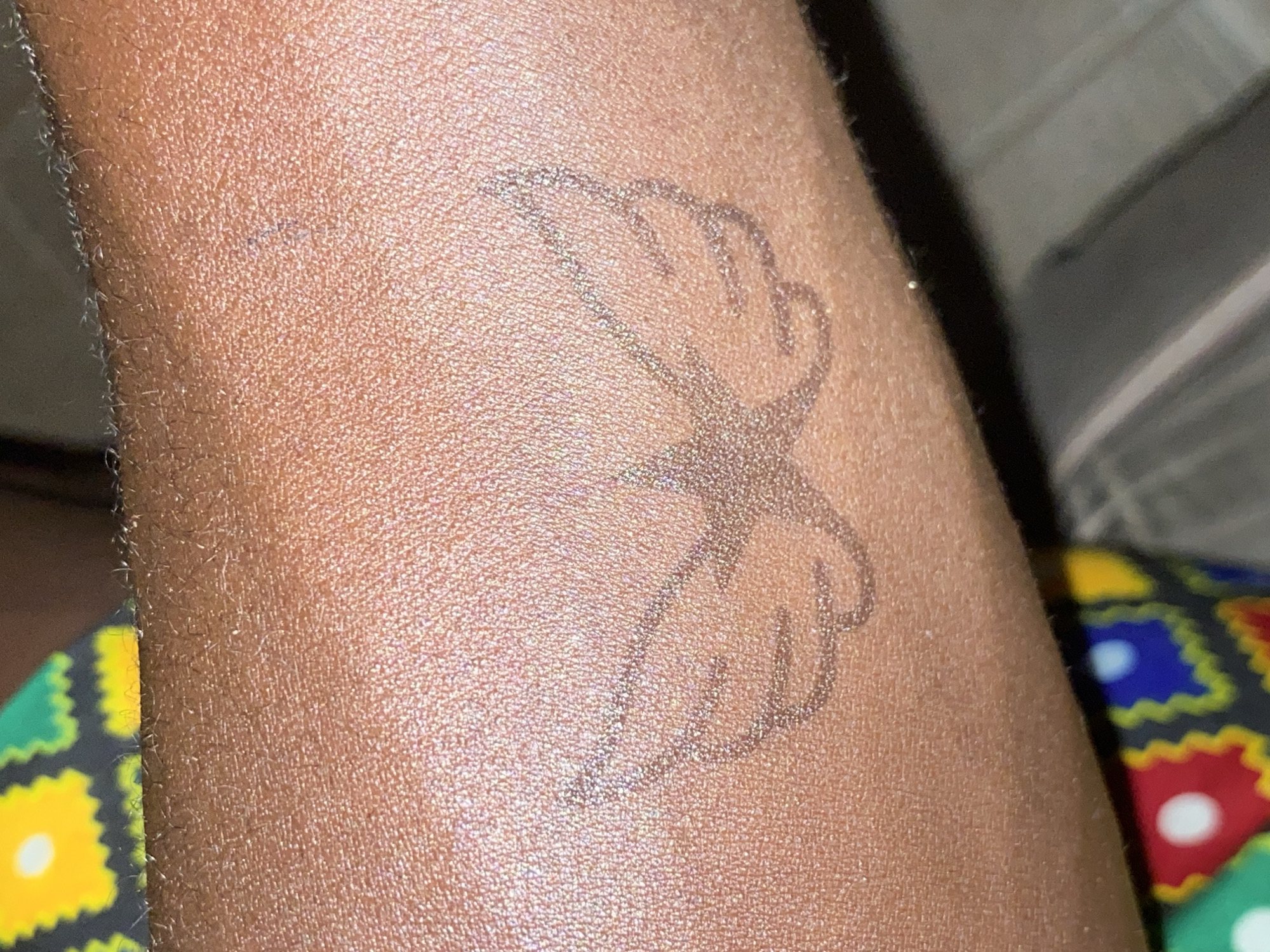A close-up image shows a person's forearm with a tattoo of a stylized design. The tattoo appears to be a butterfly shape composed of two stylized hands facing each other. The background is blurry and indistinct. The location is not discernible from the image. There is no evidence to support the subject's presence in Barden Yana, Nigeria.