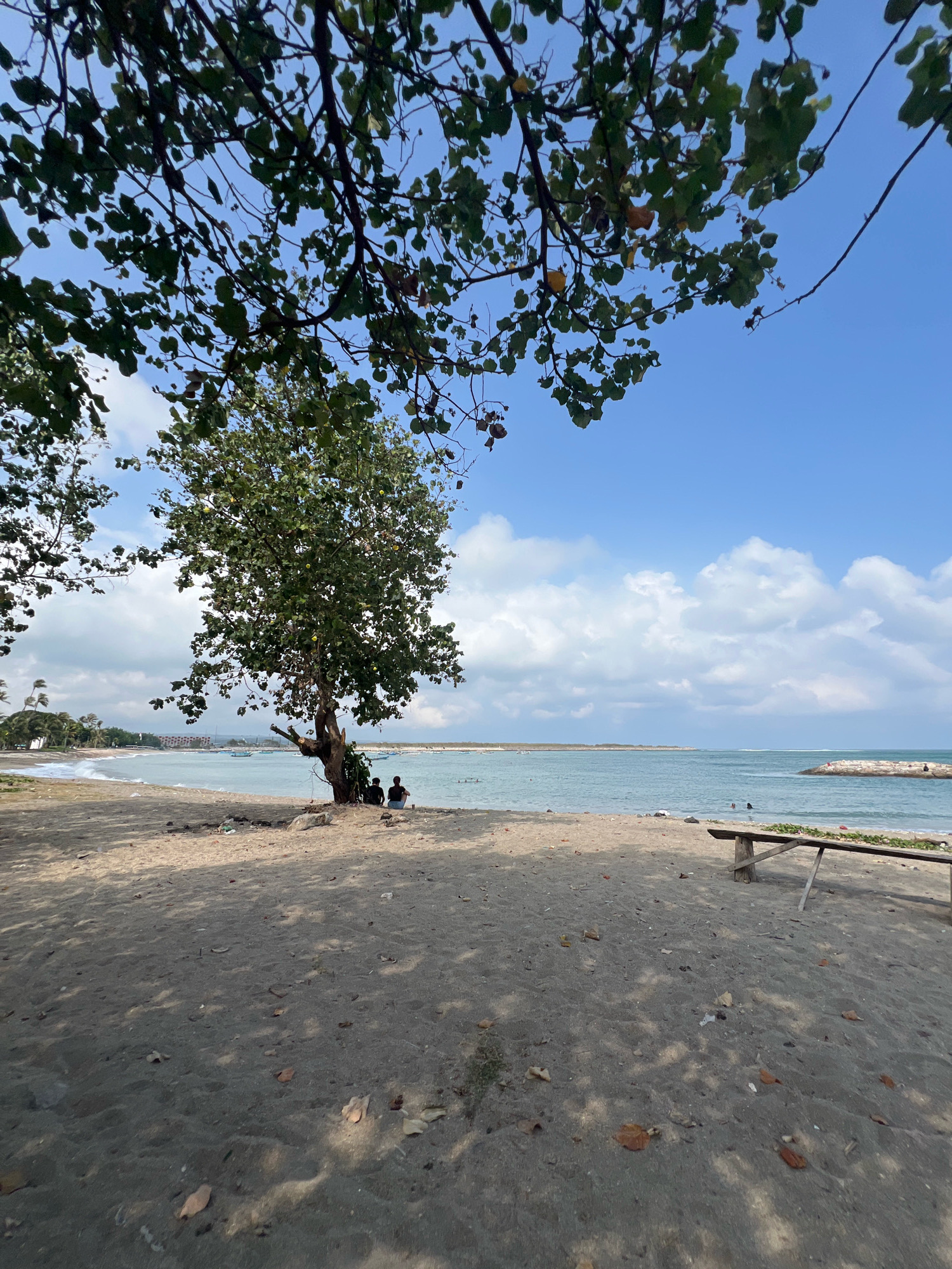 A wide-angle, low-angle shot captures a serene beach scene in Kuta, Indonesia. The foreground is dominated by dark, coarse sand scattered with dry leaves and debris, dappled with the shadows of overhead tree branches. A weathered wooden bench with a rough plank seat and diagonal supports sits in the middle ground, positioned facing the water.
Beyond the sand, the turquoise ocean stretches out, calm with small white waves breaking near the shore. In the distance, a man-made rock jetty extends into the sea, with a few figures visible on it. A lone tree with a thick, gnarled trunk and a dense canopy of green leaves stands to the left of center, providing shade over the sand. Beneath the tree, two people are seated on the sand, facing the water, appearing relaxed and engaged in conversation or contemplation.
Further back, along the coastline, palm trees and structures are visible. The sky is a vibrant blue, dotted with fluffy white clouds, suggesting fair weather. Several boats are anchored or sailing in the calm waters of the bay. The overall atmosphere is one of tranquility and natural beauty, characteristic of a tropical beach destination.