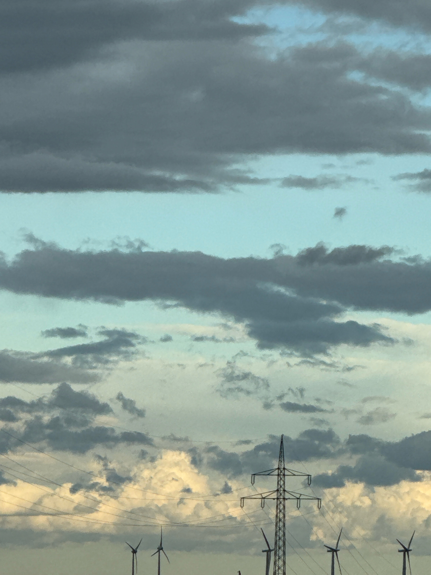 This image depicts a cloudy sky above a landscape that likely includes wind turbines and power lines, with the possibility of being near Bad Lauterberg im Harz, Germany. The sky is filled with various shades of grey and light blue clouds, suggesting a dramatic or overcast weather condition. Some clouds appear to be thick and dark, while others are thinner and more translucent, allowing hints of blue sky to show through. The lower portion of the image shows a silhouette of a power line pylon with multiple crossbars, indicative of an electricity transmission infrastructure. In the distance, the outlines of several wind turbines are visible, their tall structures and rotor blades faintly discernible against the sky. These elements collectively suggest an open, possibly rural or semi-industrial, landscape. The presence of wind turbines and power lines strongly hints at an area focused on renewable energy or industrial activity. The specific mention of Bad Lauterberg im Harz, Germany, suggests a context for this scene, possibly indicating a region known for its wind farms or power infrastructure. No people are visible in the image. The overall scene is static, with no discernible actions or interactions. The lighting is diffused due to the cloud cover, creating a somewhat muted color palette.