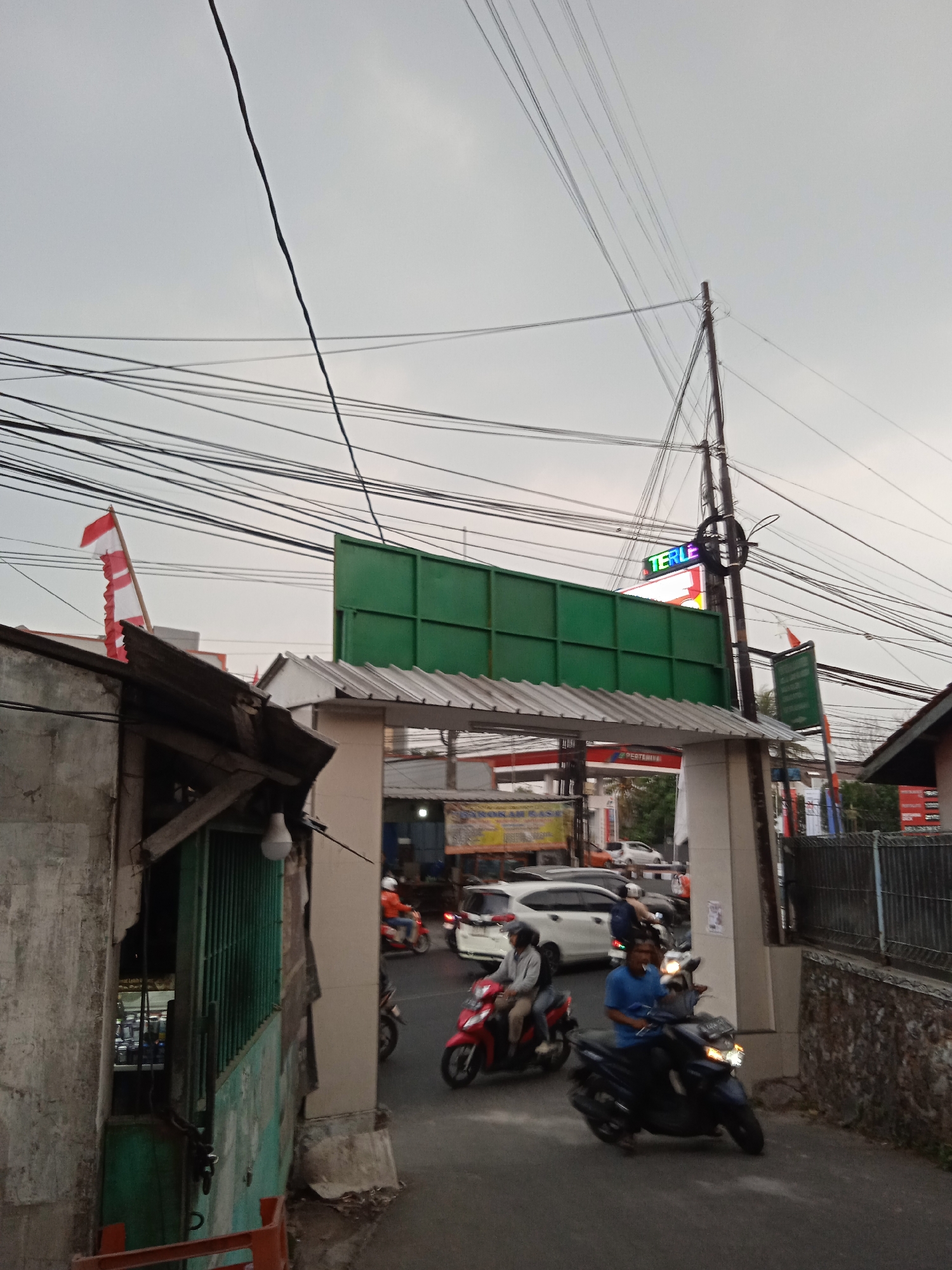 A low-angle, vertical, medium shot captures a street scene in Cileunyi, Indonesia, under an overcast sky. Numerous black electrical wires crisscross the frame horizontally and diagonally, supported by a prominent utility pole on the right. In the foreground, on the left, is a weathered, grey concrete wall with a dilapidated wooden structure extending from it, featuring green barred windows. To the right of this, an arched gateway leads to a road where several motorbikes and a white car are in motion. Two men are riding motorbikes in the foreground; one on a red motorbike, and another on a dark blue motorbike. The man on the blue motorbike is wearing a blue t-shirt, and the man on the red motorbike is wearing a white t-shirt. Behind them, a white car is visible. In the background, a green billboard stands above the gateway. A red and white flag is flying on a pole to the left of the billboard. A sign with "TERLE" illuminated in colorful LED lights is attached to a utility pole. Further back, a Pertamina gas station is visible with its red and white signage. The scene appears to be a busy street with active traffic and ongoing daily life.