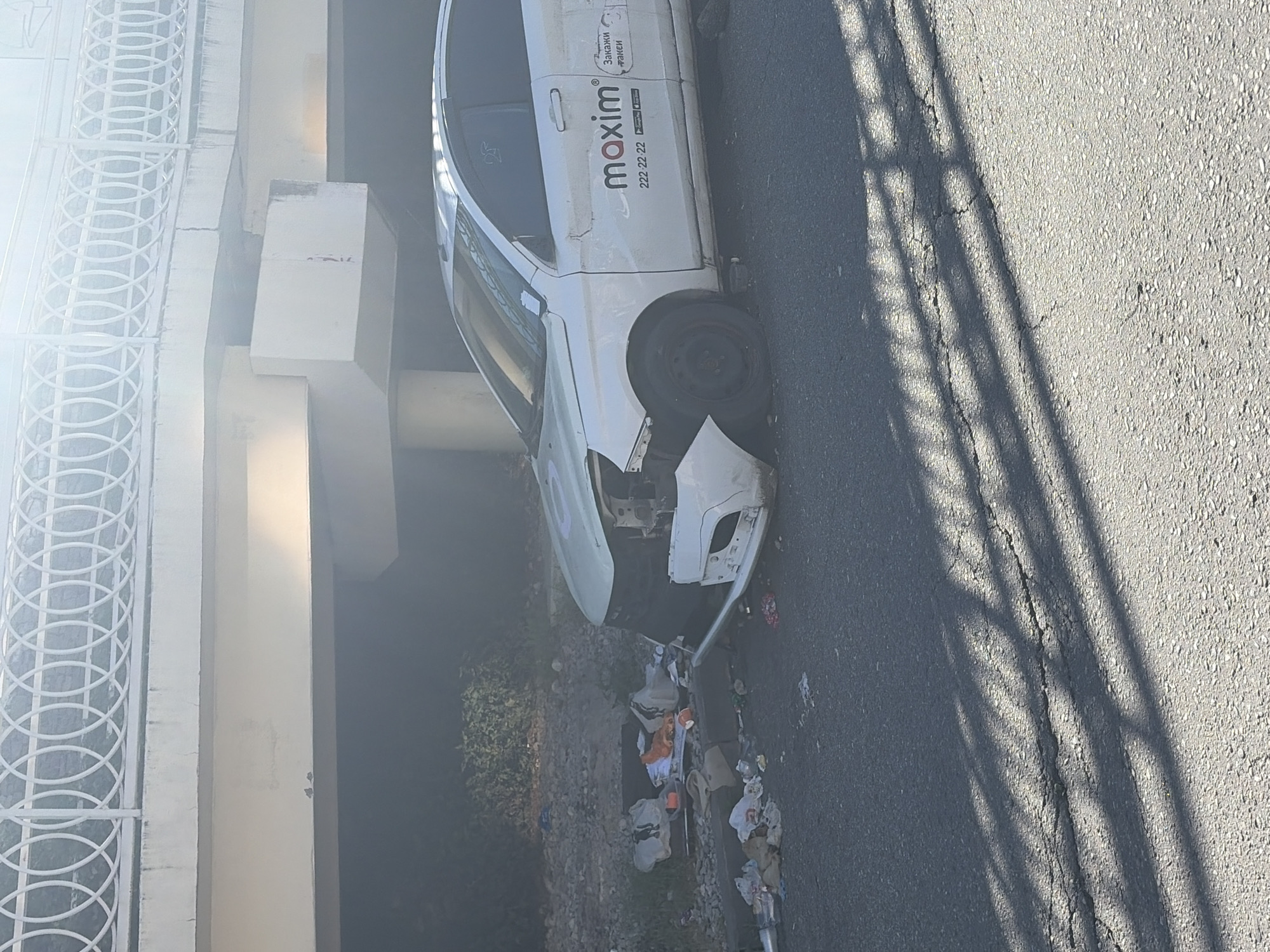 This media file captures the aftermath of a car accident under a concrete overpass or bridge structure in Sochi, Russia, during daytime. The primary subject is a white car, identified as a "maxim" taxi through branding on its side door, which includes the company name, phone number "222-22-22," and the Cyrillic text "Закажи такси" (Order a taxi), along with app store logos.
The car has sustained significant damage to its front end; the bumper is detached, and the hood is severely crumpled, exposing internal components. It is positioned off the asphalt road, perpendicular to the road's flow, near a concrete barrier. Adjacent to the car and barrier, a considerable amount of debris and trash, including plastic bags and bottles, is scattered on the ground.
Bright sunlight is evident, casting long, distinct shadows, possibly from another fence or structure, across the cracked asphalt road. To the left, a white metal fence with circular patterns, part of the bridge railing, and massive concrete support pillars of the overpass are visible. Green foliage can be seen beneath the bridge structure in the background. No people are present in the image.