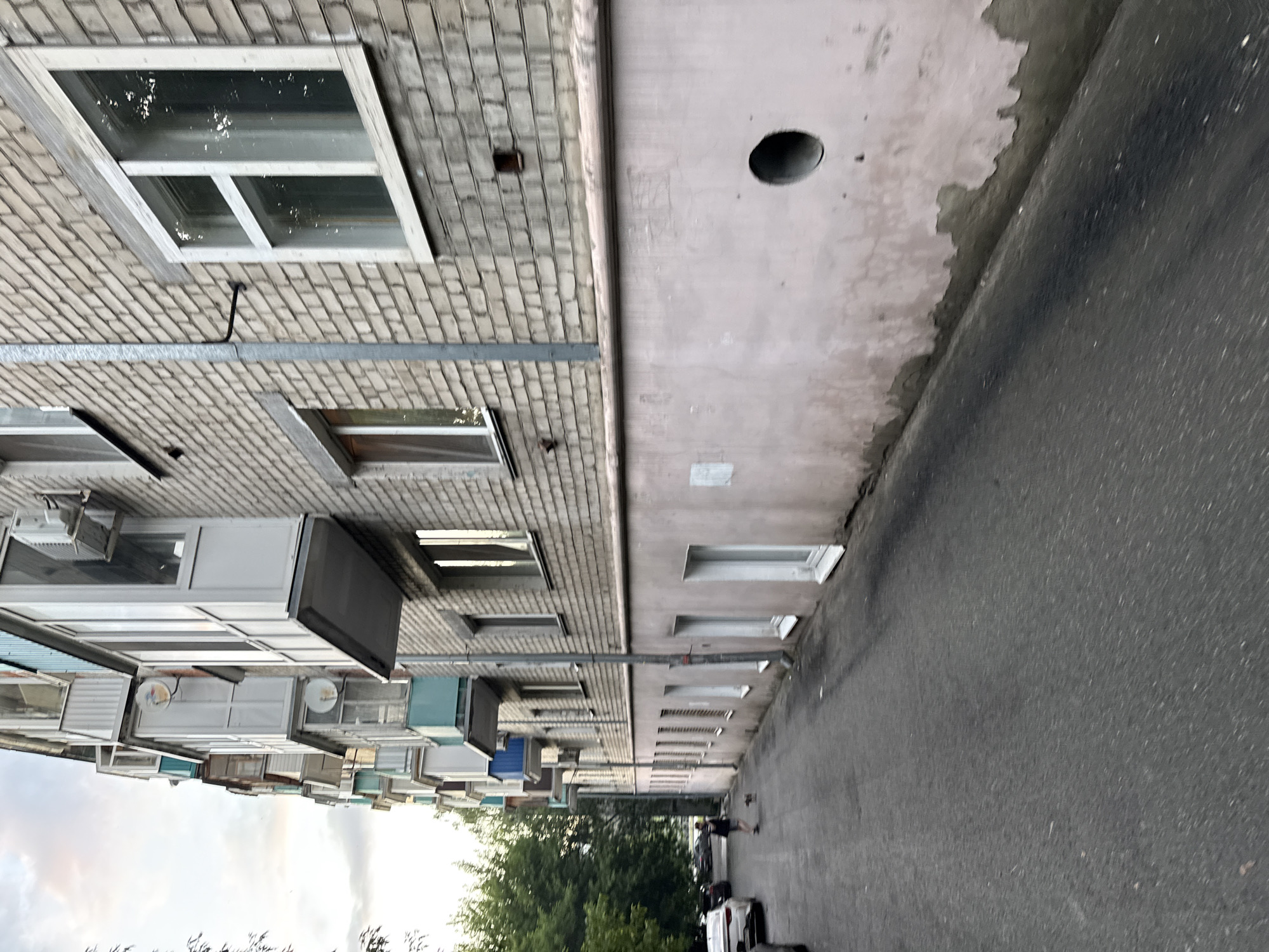 The image depicts an exterior view of a multi-story apartment building in Kazan, Russia. The building's facade is constructed of light-colored bricks, with several balconies extending from the upper floors, some of which are enclosed with glass. The lower portion of the building features a light pink, weathered wall with several windows and a large circular opening. An asphalt pathway runs alongside the building, and a person is visible in the distance walking a small dog. Trees and parked cars are also present, indicating an urban residential setting. The sky is overcast with clouds.
