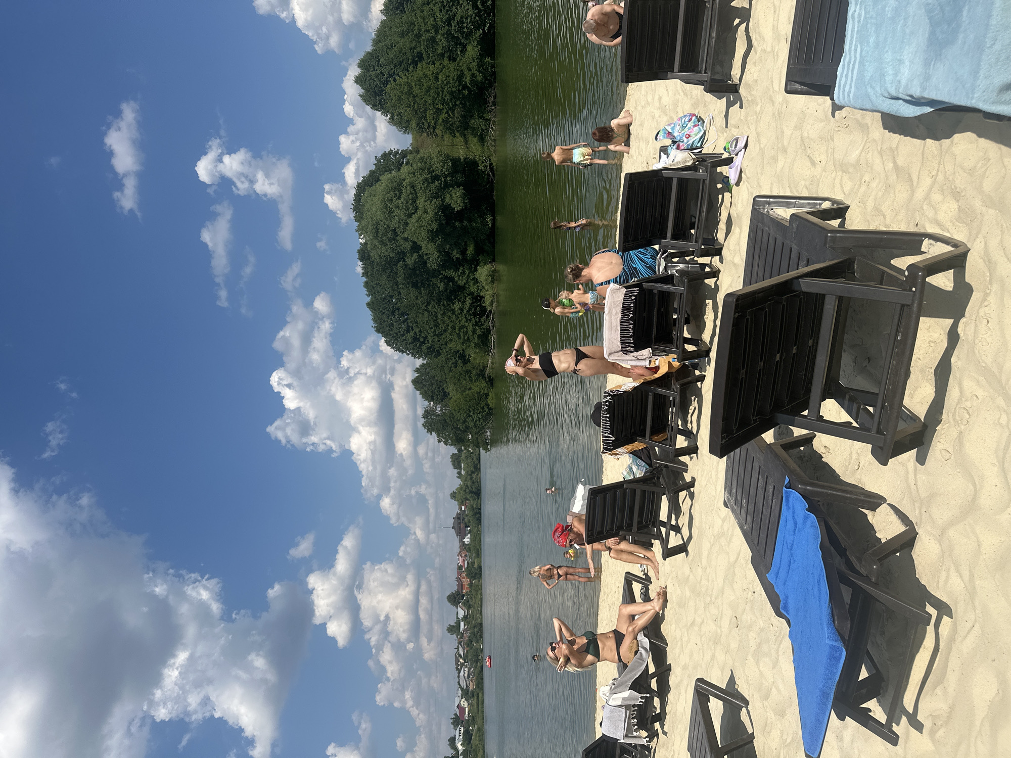 The image depicts a sunny day at a sandy beach next to a body of water, possibly a lake or river, with lush green trees and a village visible in the background. The sky is a vibrant blue with fluffy white clouds. Several people are present, some sunbathing on black lounge chairs scattered on the sand, while others are in the water.
In the foreground, a woman in a dark bikini is lying on a lounge chair, facing the sun, with her hands resting on her head. Another woman in a green bikini is also sunbathing nearby. To the left, a woman in a black bikini stands with her hands on her hips, looking towards the water. Further back, several individuals are visible in the water, some appearing to swim or wade. A man in patterned swim trunks is standing at the edge of the water.
Various objects are present on the sand, including beach towels of different colors, a patterned beach bag, and flip-flops. The black lounge chairs are a prominent feature of the scene, suggesting a recreational area. The overall atmosphere appears relaxed and leisurely, consistent with a warm summer day at a beach. The presence of the village and dense trees indicates a natural, possibly semi-rural setting. The time of day is likely midday or early afternoon, given the bright sunlight and shadows. No visible text is present in the image.