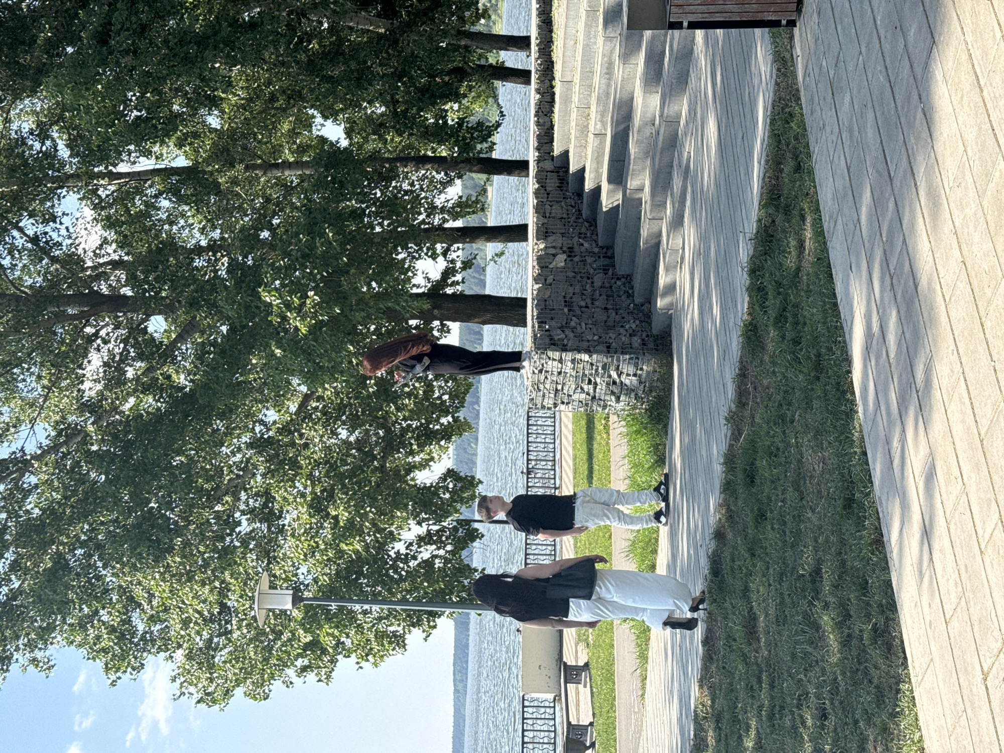 The image depicts a scene in Votkinsk, Russia, near a body of water, likely the Votkinsk Reservoir. The view is from a pathway bordering the water. On the left, there are mature trees with dense foliage. A street lamp is visible among the trees. People are present on the pathway. Two people, a man and a woman, are near the bottom of the frame. The woman has dark hair and is wearing dark clothing on top and light-colored pants. The man is wearing a dark top and light-colored pants. Further up the path, another woman with long brown hair and dark pants is standing near a rock retaining wall that forms a transition to a set of stairs. The pathway is paved with light-colored stones and there is a strip of grass between the walkway and the building in the right. The water in the background is calm. The sky is light blue with some clouds visible.