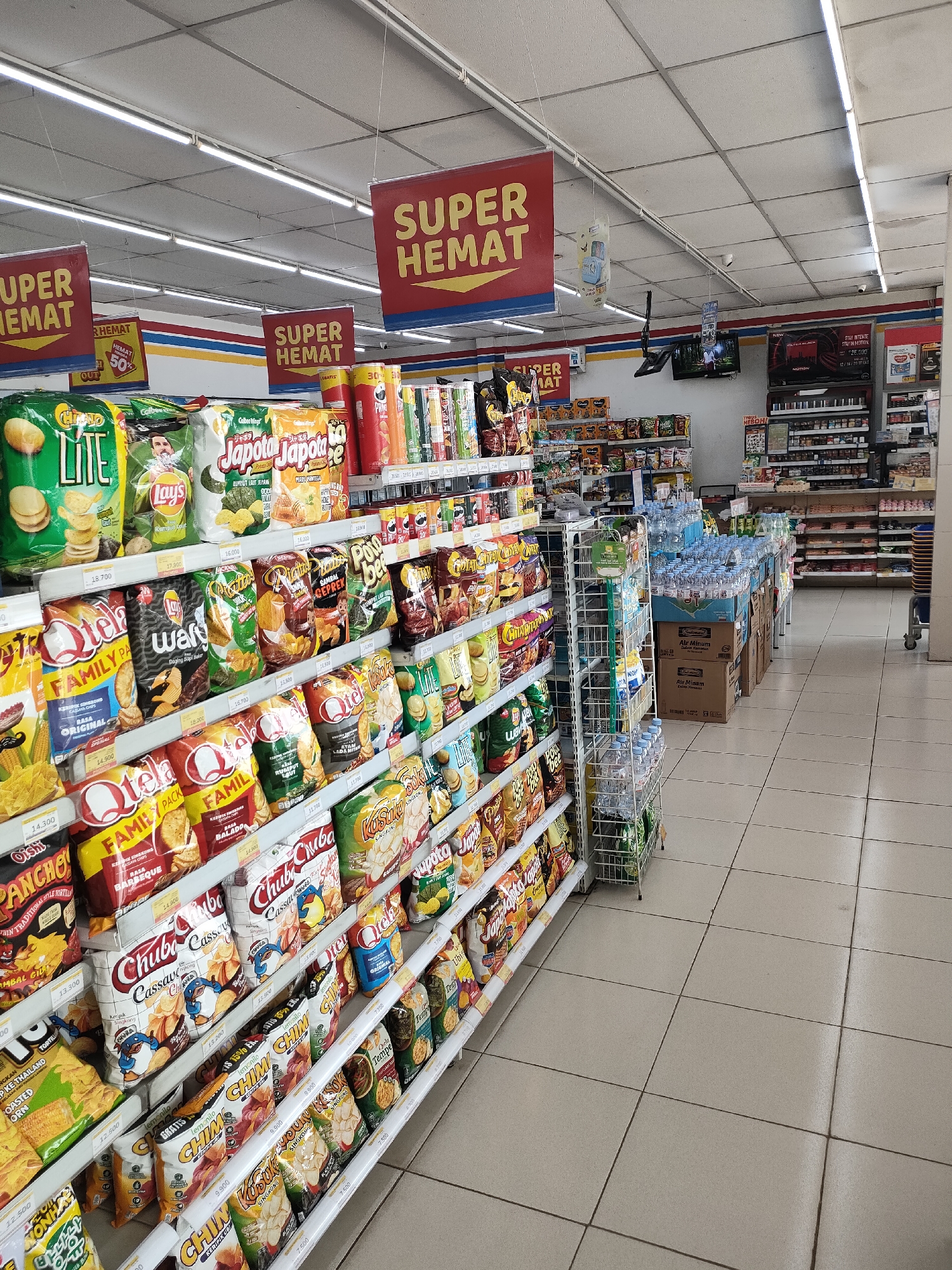 The image depicts the interior of a convenience store, specifically a Minimarket, in Curahdringu, Indonesia. The scene is dominated by brightly lit aisles filled with shelves stocked with various food items, primarily snacks and beverages. Several prominent red and yellow signs with the text "SUPER HEMAT" are hung from the ceiling, indicating promotional sales.
In the foreground, shelves are packed with an assortment of bagged snacks, including chips like Lay's, Chiki Family, and various other brands with Indonesian text. Some of these bags are colorful and feature images of food or cartoon characters. Adjacent to the snack aisles, there is a display of bottled water. Further into the store, more shelves are visible, holding a wider range of products, including what appear to be canned goods or other packaged food items. Behind these shelves, a section of the store shows more organized shelving, possibly for household items or toiletries.
The flooring is tiled in a light beige color, with visible grout lines. The ceiling features fluorescent lighting fixtures and a tiled pattern. In the background, a television screen is mounted on the wall, displaying an image. The store appears to be well-stocked and organized, typical of a retail environment. No individuals are clearly visible in the image, suggesting the photograph was taken when the store was either empty or people were out of frame. The overall impression is one of a modern, functional convenience store offering a variety of consumer goods.