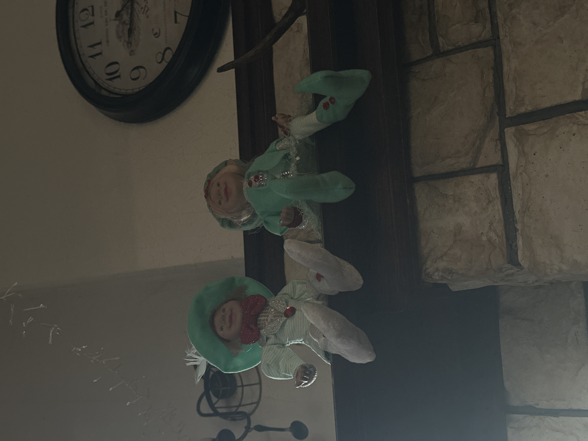 The image shows a room interior with several elements. A clock with Roman numerals is mounted on a wall. Two elf dolls, dressed in green and white outfits, are positioned vertically against a dark surface, likely a door or a piece of furniture. A stone wall is visible on the right side of the image. A black wrought iron decorative item is on the left side of the image.